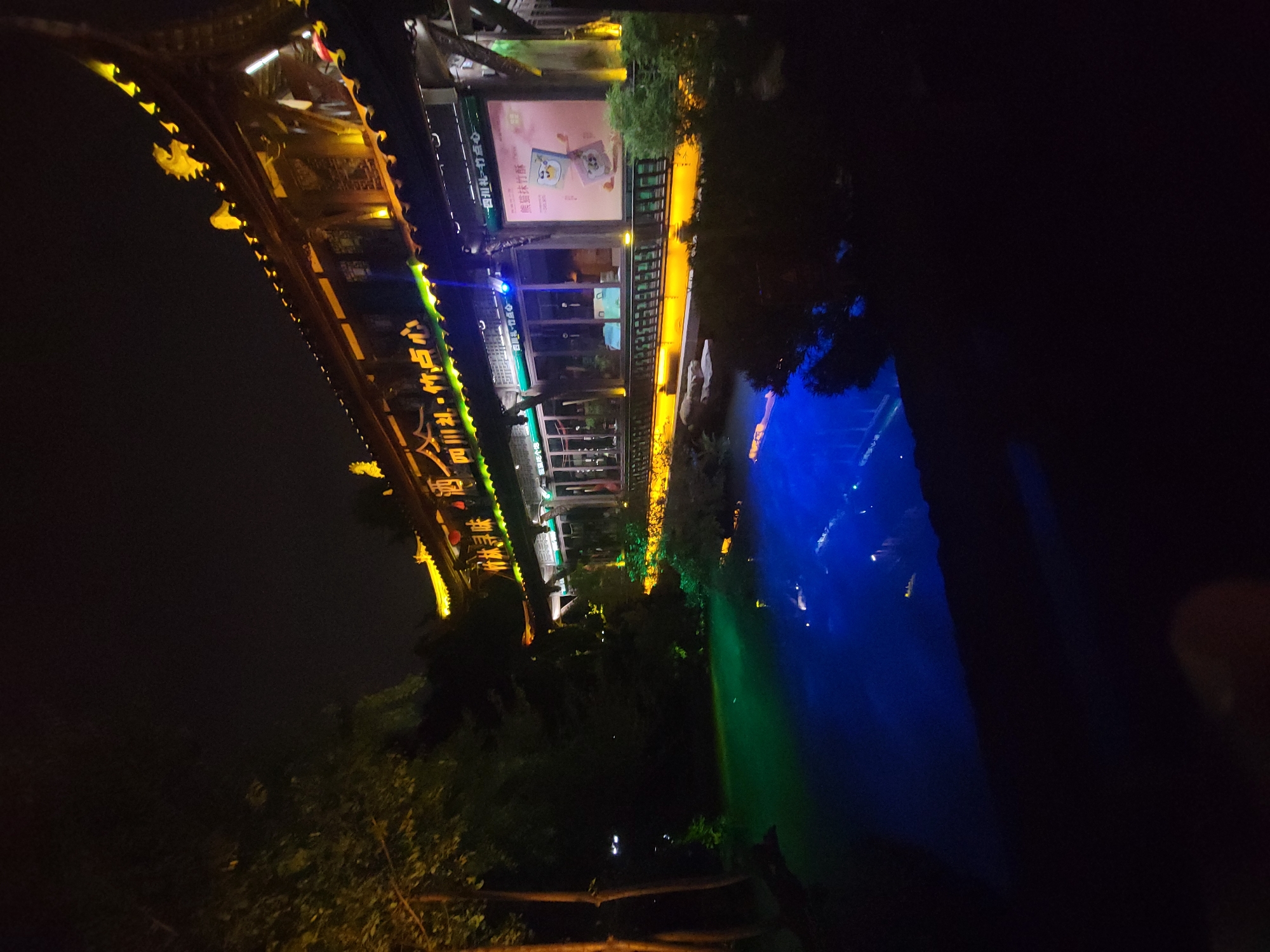 A traditional Chinese restaurant building is illuminated at night, overlooking a body of water that reflects the building's lights. The building features ornate architecture with a dark, tiled roof and yellow decorative elements, including a dragon motif. Neon lights, primarily green and blue, illuminate the signage and the exterior of the building. A poster with cartoon characters is visible on the building's facade. The water in front of the building is lit with alternating green and blue lights, creating a vibrant and surreal effect. Trees are visible to the left of the building, their leaves catching some of the yellow light. The scene appears to be in a park or recreational area within Wuhou District, China, likely a tourist attraction or a popular dining spot. No people are visible in the image.