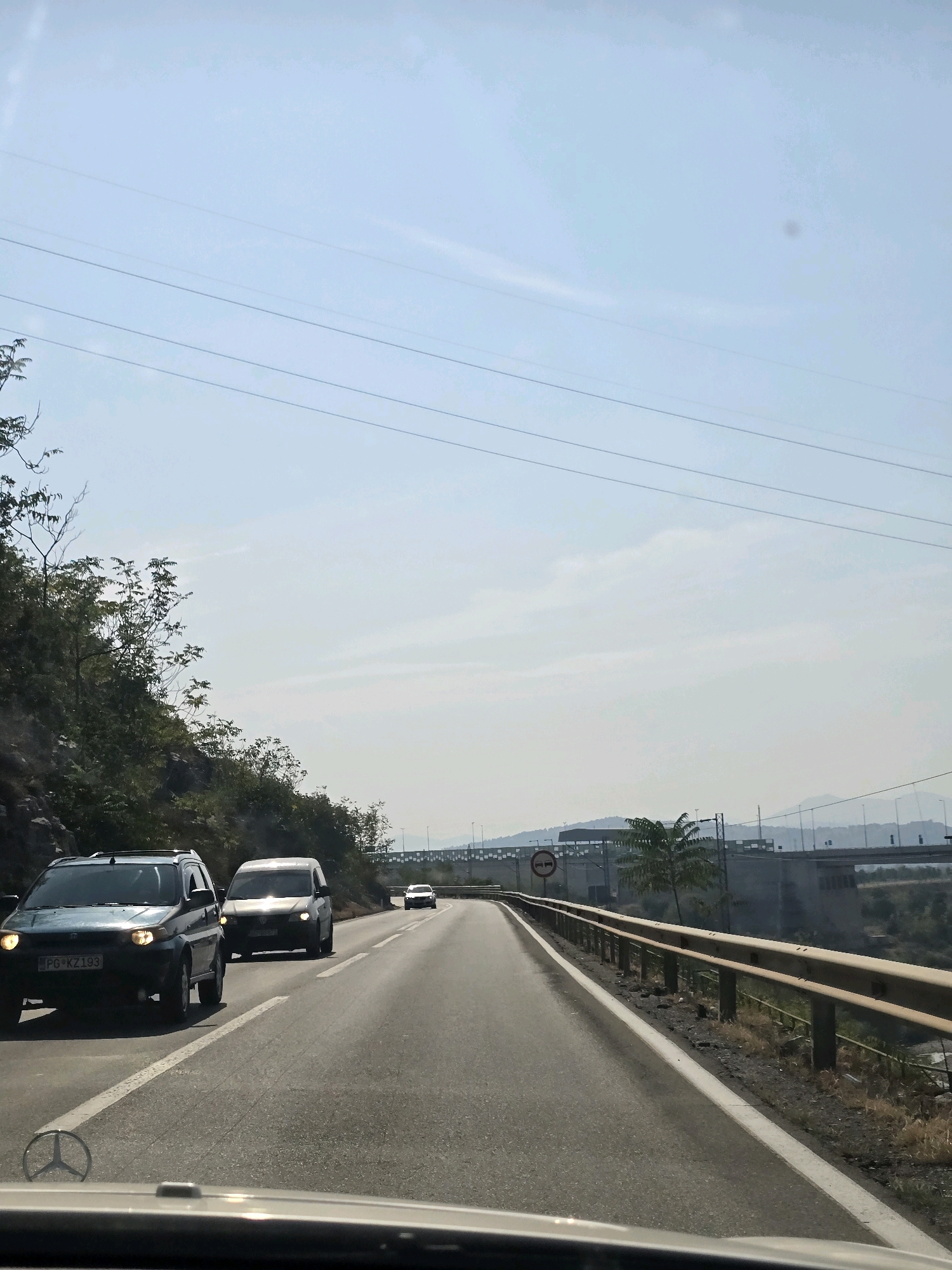 A view from inside a vehicle traveling on a highway in Montenegro, likely near Podgorica, as indicated by the license plate "PG KZ 193" on the dark blue SUV in the foreground. The highway has two lanes in the direction of travel and is bordered by a guardrail and a steep, vegetated hillside on the left. Several vehicles are visible on the road, including the dark blue SUV and a silver minivan in front of it, both with headlights on. Further ahead, another white vehicle is visible in the distance. The sky is clear with a few wispy clouds, and power lines stretch across the frame. In the background, there is evidence of infrastructure such as a bridge or overpass and distant hills. The overall scene depicts a daytime journey on a scenic road.