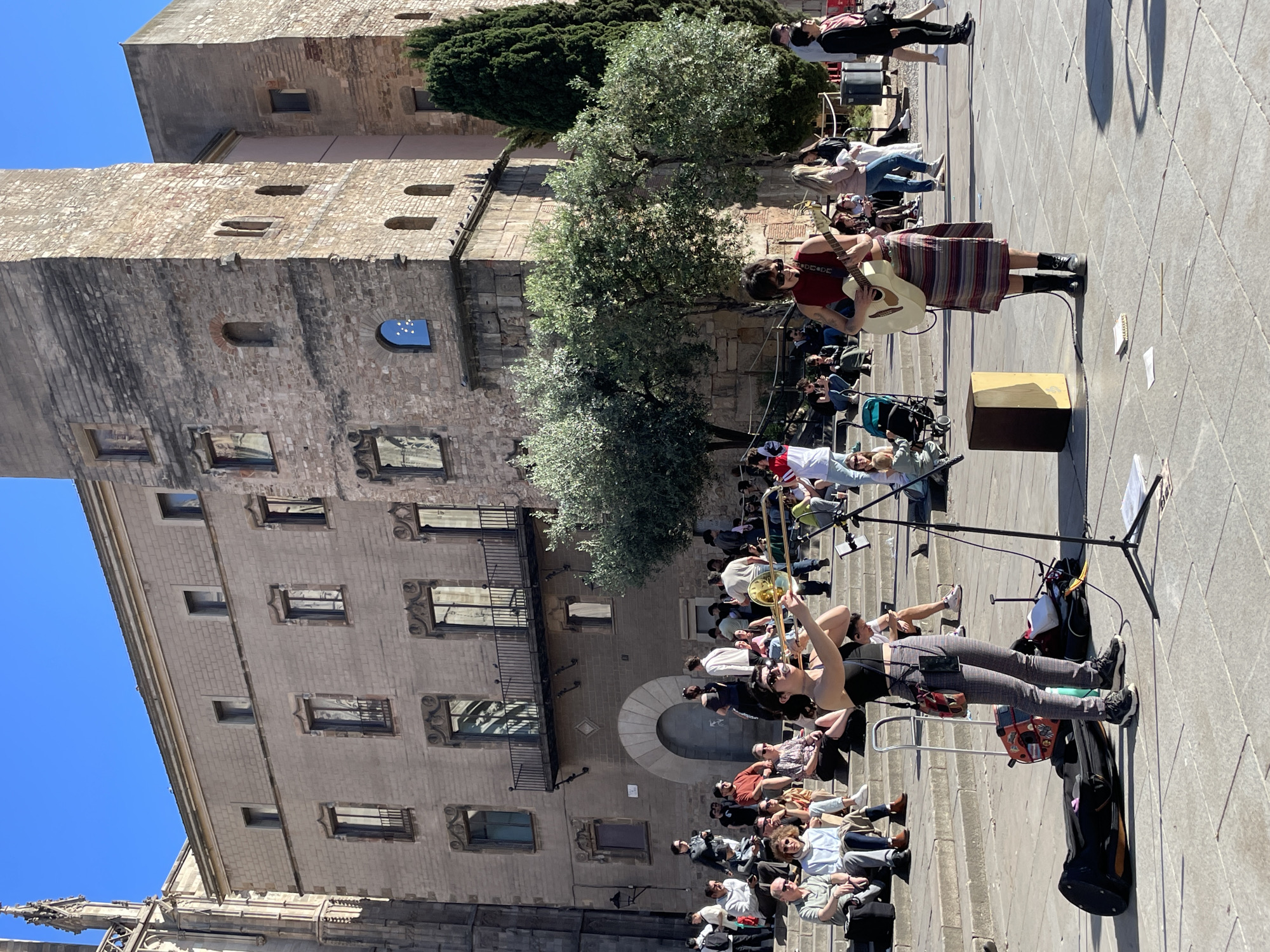 A street scene in Barcelona, Spain, shows two musicians performing on a public square, with a large stone building in the background. The building has multiple windows, a balcony, and a tower-like structure. The musicians are in the foreground, facing a seated audience on steps. One musician plays a trombone, and the other plays a guitar and sings into a microphone. A gold-colored box sits near the guitar player. People of varying ages and demographics are sitting or standing, watching the performance. A bright blue sky is visible above the building. The scene suggests a live music performance in an outdoor public space, likely a tourist destination in Barcelona.