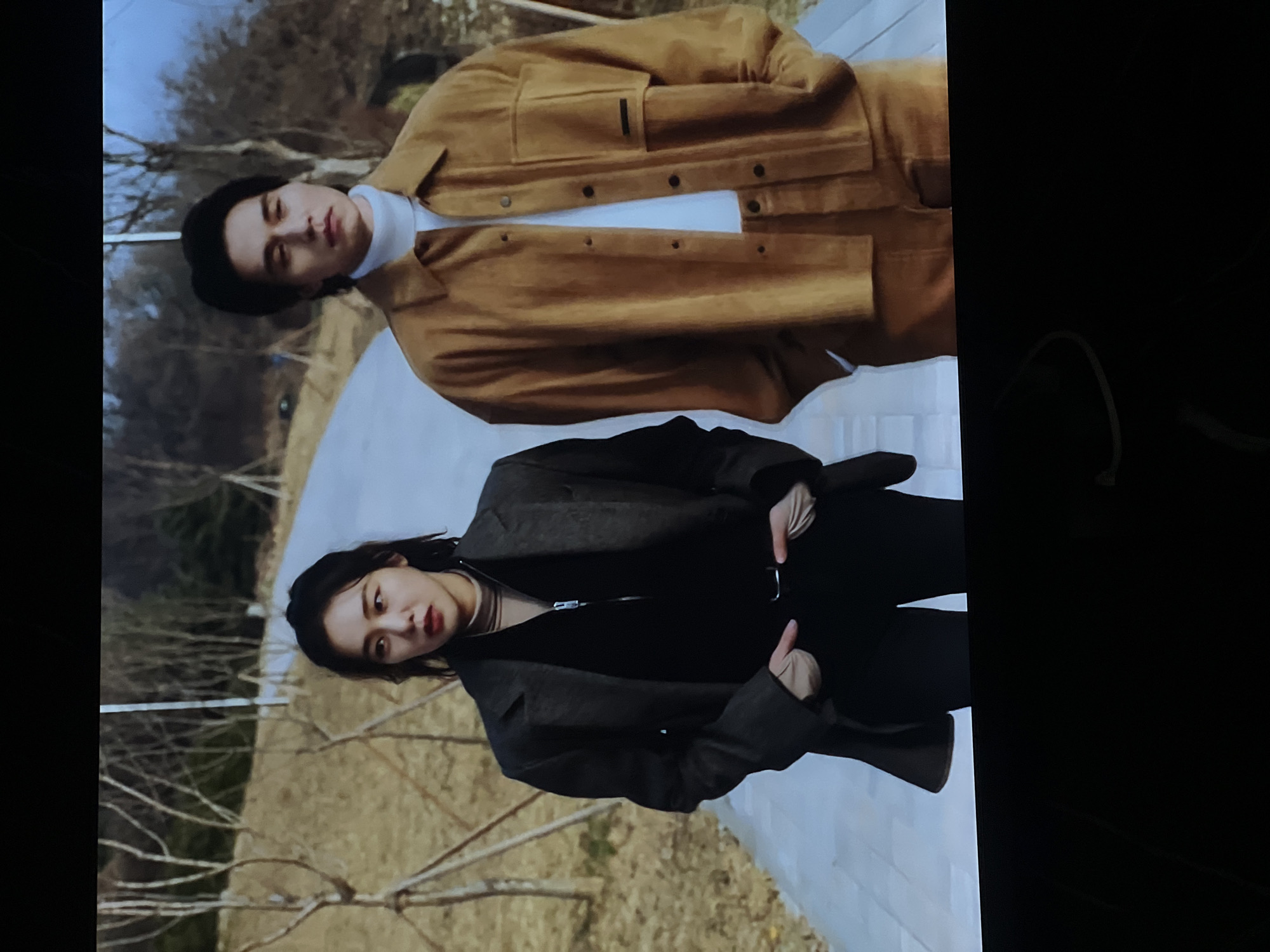 Two people stand outside on a stone path in a park-like area. A man wears a black cap, a white turtleneck sweater, and a brown jacket. A woman wears a black coat and pants. The background includes trees and a cloudy sky. The location is likely not Vologda, Russia, as the environment does not match the typical landscapes and architectural styles associated with the city.
