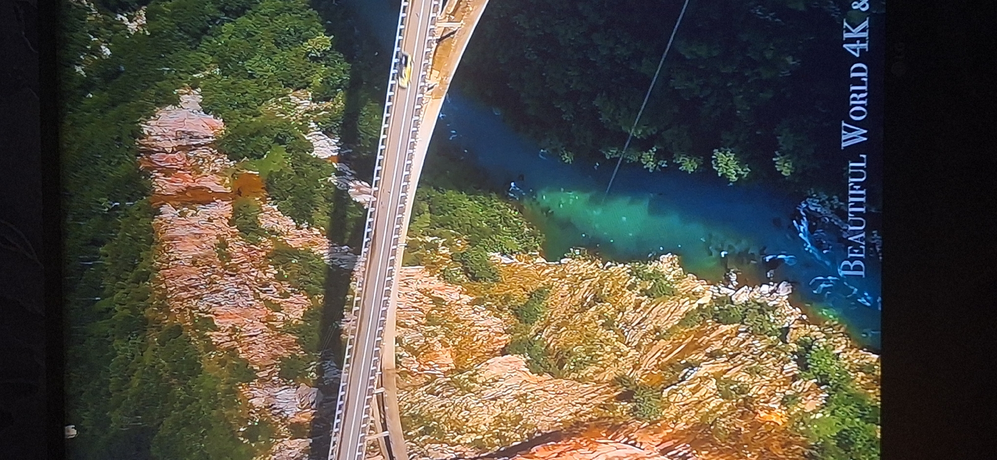 The image shows an aerial view of a deep gorge with a river flowing through it. A modern bridge spans the gorge, carrying a road with at least one yellow car. The sides of the gorge are steep cliffs covered in vegetation. The water in the river is a striking turquoise color. At the bottom of the image, white text reads "BEAUTIFUL WORLD 4K &".
There is no information in the image to suggest this location is in Bila Tserkva, Ukraine. The landscape depicted is more characteristic of a mountainous or canyon region.