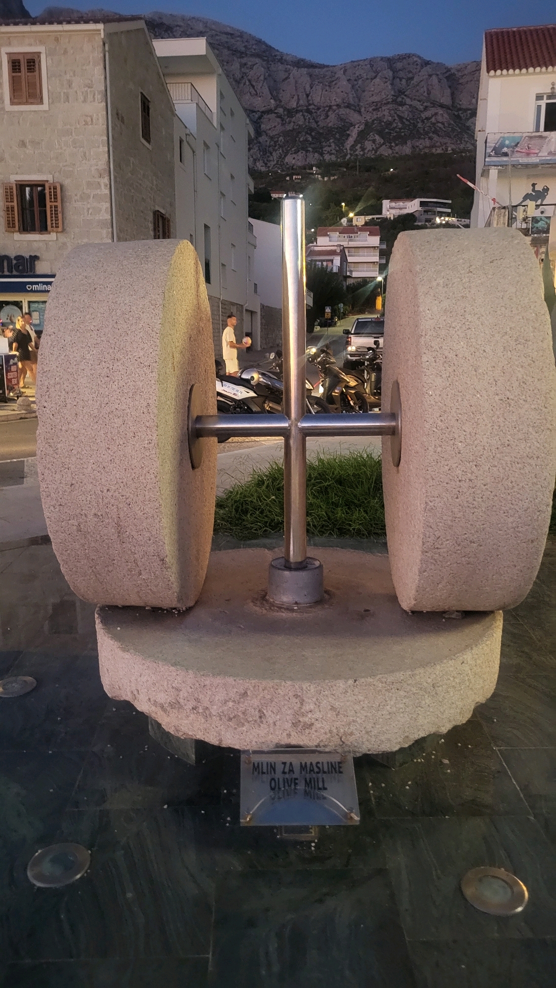 The image shows an outdoor scene in Općina Tučepi, Croatia, at dusk. The central subject is a large, circular stone olive mill, with two massive millstones positioned vertically and facing each other. A metal pole runs vertically through the center of the millstones, securing them to a circular stone base. In front of the millstones, on the base, is a small, rectangular sign with text that reads "MLIN ZA MASLINE OLIVE MILL". The mill is situated on a dark, reflective tiled surface.
In the background, there are buildings, suggesting an urban or town setting. To the left, a light-colored building with stone accents and wooden shutters is visible. Further back, a modern white building with balconies stands against a backdrop of a large, rocky mountain range. The sky is a deep blue, indicating the transition from daylight to night.
To the left of the olive mill, a few people are present. One person, a man in a white t-shirt and shorts, is standing and holding a ball. Another person, a woman in dark clothing, is walking away from the camera. Several motorcycles are parked behind the mill. The overall impression is a public space, likely a tourist or town square, featuring a historical artifact or monument.