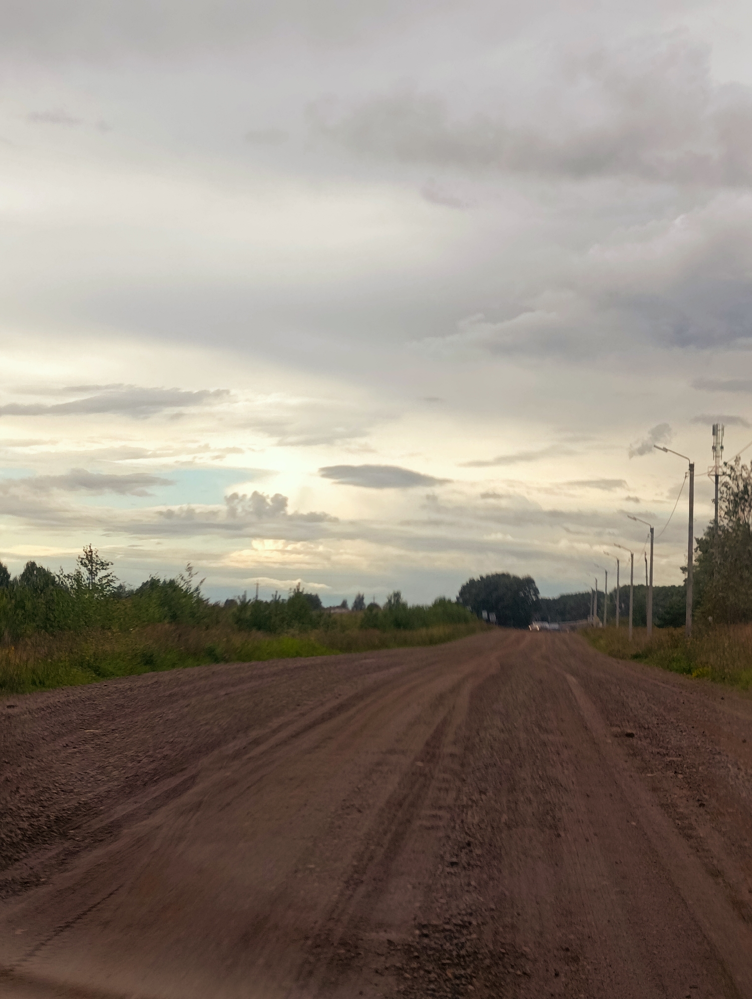 This is a low-angle, eye-level, full shot of a dirt road in Kamennyi Kliuch, Russia, with a cloudy sky overhead. The road, which appears to be unpaved, takes up the majority of the foreground and middle ground, showing tire tracks and a rough surface composed of dirt and gravel. On either side of the road, there is tall grass and vegetation, with trees and bushes further in the distance. Several utility poles with lights and what appears to be a cell tower are visible along the right side of the road. In the far distance, there are faint lights suggesting vehicles or buildings. The overall lighting suggests it is either dawn or dusk, with the sun partially visible through the clouds.