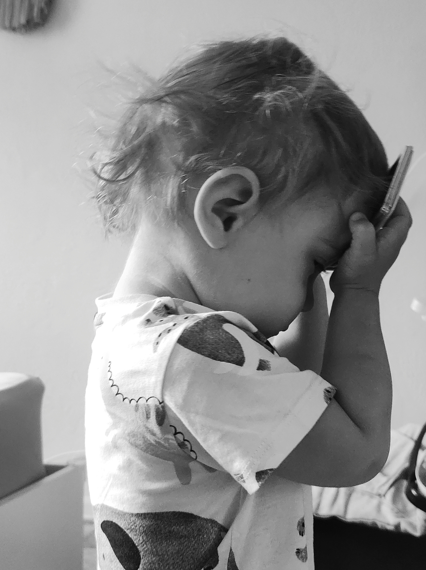 A close-up, black and white portrait features a young child, likely a toddler or preschooler, captured in a moment of deep thought or contemplation. The child is seen in profile from the right side, with their head bowed and eyes cast downwards or closed. Their hands are cupped around their face and temple, with a thin, elongated object, possibly a pen or a small toy, held against their forehead.
The child has light-colored, slightly disheveled hair. They are wearing a short-sleeved t-shirt adorned with a light-on-dark pattern that appears to include stylized animal outlines, perhaps dinosaurs, and wavy, squiggly lines. The fabric of the shirt has visible texture.
The background is simple and uncluttered, primarily consisting of a plain, light-toned wall, suggesting an indoor setting. On the far left, a blurry, lighter object is partially visible, likely a piece of furniture. To the right, a darker, out-of-focus shape can be seen.
The posture of the child conveys a sense of intense focus, concentration, or possibly weariness. There are no overt signs of distress, but rather an introspective mood. No specific time of day or external weather conditions can be discerned from the image. There is no visible text. The scene is located in Chaikovsky, Russia.