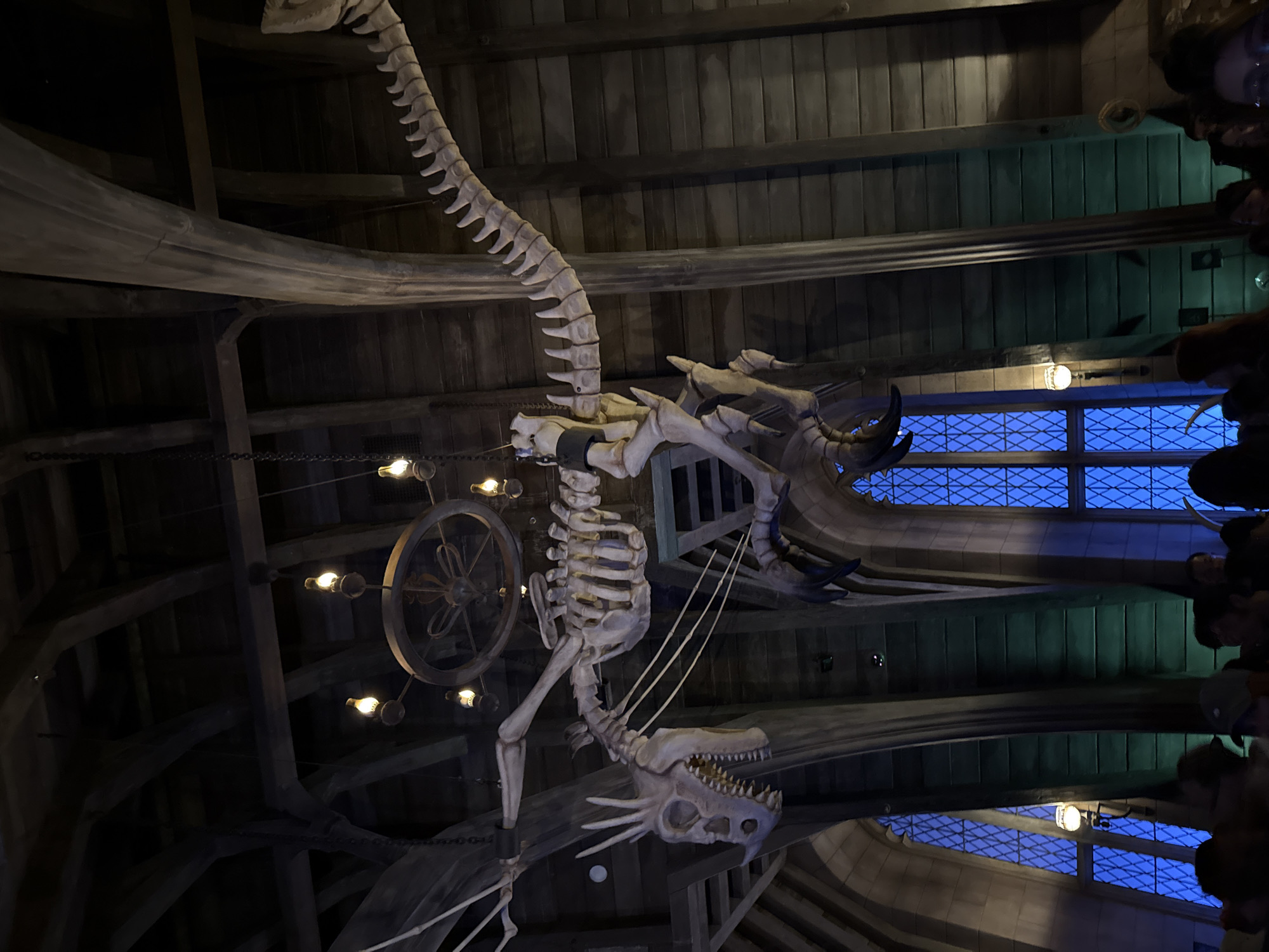 The image depicts the interior of a building, likely a restaurant or themed attraction. A large, skeletal dragon structure hangs from the wooden-beamed ceiling. To the left of the dragon is a circular light fixture with several lightbulbs. In the background, a window with a blue-tinted grid is visible. On the right side, a dark mass of people is partially visible, their forms indistinct. The style suggests a gothic or fantasy setting. There is no specific indication of the location being in Tongzhou District, China.