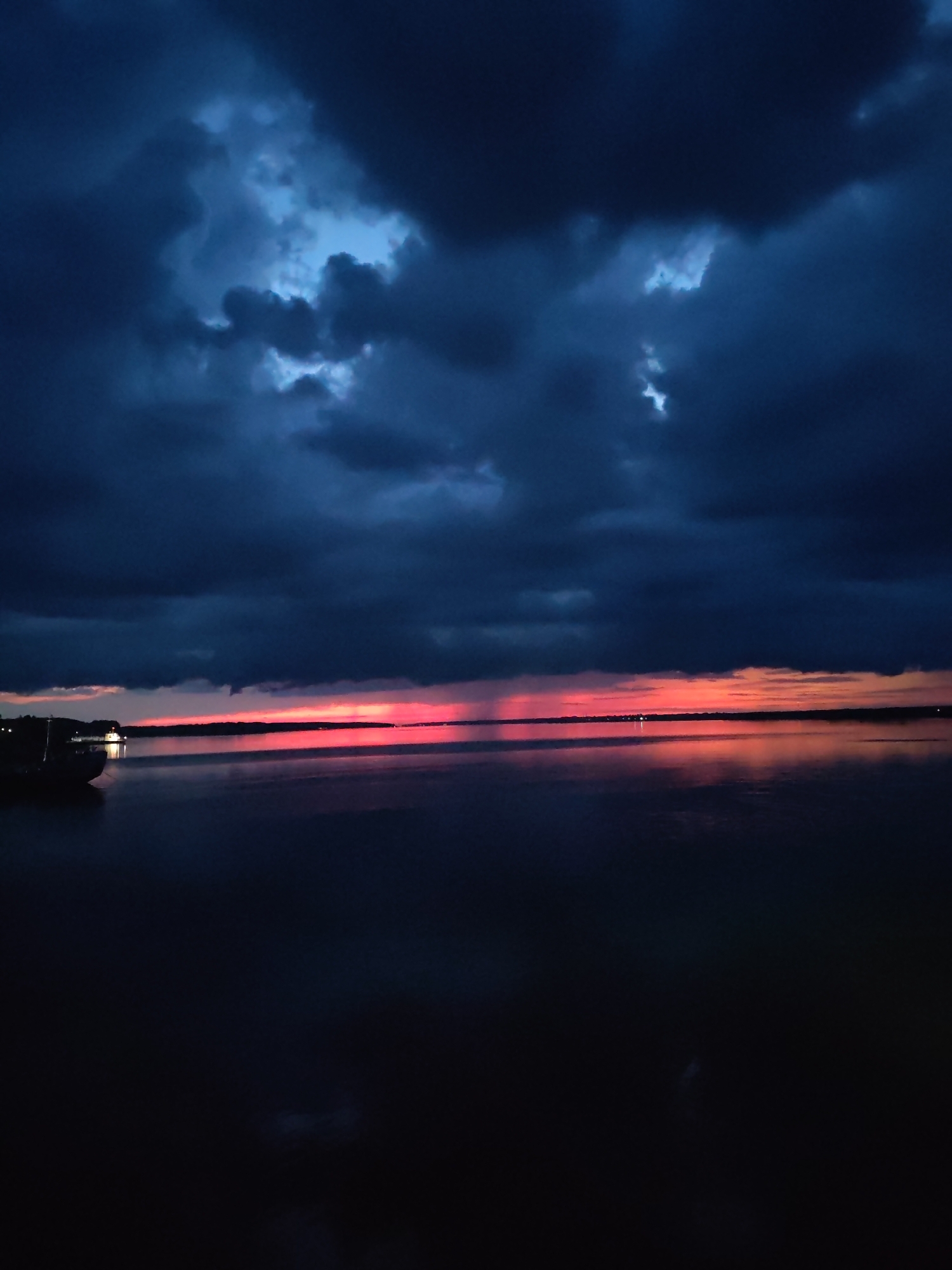 A body of water, possibly a river or lake, is depicted under a stormy, twilight sky. Dark, heavy clouds dominate the upper portion of the image, with patches of lighter blue sky visible through breaks in the clouds. A vibrant band of pink and orange light stretches across the horizon, reflecting on the calm water. On the left side of the image, a dark silhouette of a boat or dock is visible near the water's edge, with a small, illuminated structure on it. The overall mood is dramatic and serene, with the contrast between the dark sky and the colorful sunset. This scene could be located along the Volga River in Rybinsk, Russia, as the city is situated on the river and known for its scenic waterfronts.