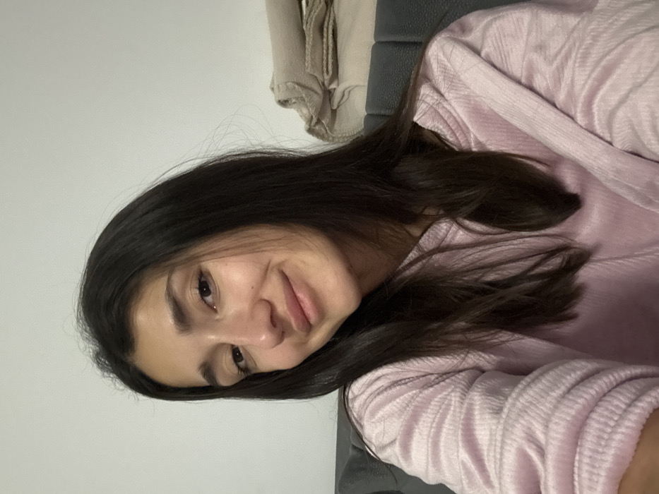The image displays a head-and-shoulders portrait of a young woman, captured in what appears to be a selfie or close-up shot. The image is oriented sideways, suggesting it was taken either by the subject lying down or the phone was held in a landscape orientation and then rotated.
The woman has long, dark brown or black hair that falls over her shoulders and chest. Her skin tone is fair to medium, and her complexion appears smooth. She is looking directly at the camera with dark eyes and a gentle, closed-mouth smile. Her eyebrows are well-defined and dark, matching her hair color. A slight crinkle around her eyes and a subtle dimple on her left cheek are visible, contributing to her friendly expression. Her head is tilted slightly.
She is wearing a long-sleeved top in a light pink or possibly very pale lavender color. The fabric has a distinct ribbed or textured pattern, particularly visible on the sleeve and across her torso.
The background consists of two main areas. To the upper left (when viewed in its current rotated state), there is a plain, light-colored wall, likely white or off-white. To the upper right and directly behind her, a dark gray or charcoal-colored cushioned surface is visible, possibly part of a couch, chair, or headboard. Above this dark surface, some folded neutral-colored fabrics, perhaps blankets or clothing items in beige or light brown tones, are casually placed.
The lighting is soft and even, suggesting an indoor setting, and there are no harsh shadows, illuminating her face clearly. The overall mood of the image is calm and pleasant. No text is visible in the image.