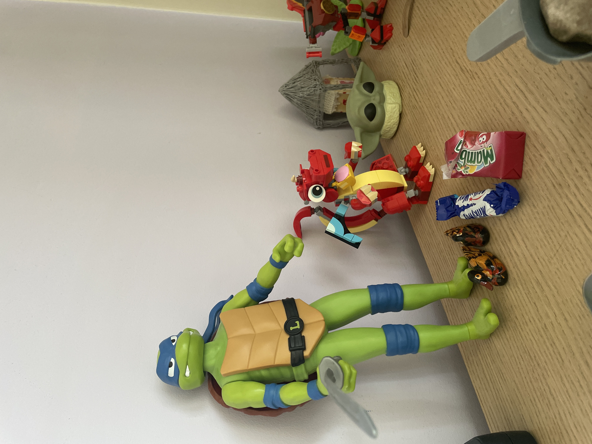 In an indoor setting, likely a shelf or table, several toys are arranged. Dominating the left side is a large, green Teenage Mutant Ninja Turtle action figure, identified as Leonardo by the "L" on his belt buckle. He is holding a silver sword in his right hand. To his right, a red Lego dragon figure with yellow accents and large googly eyes stands proudly. Behind the dragon, a small green figure resembling Grogu from The Mandalorian, also known as Baby Yoda, sits on a light green base. Further to the right, a red and orange Lego robot is partially visible. In the foreground, near the bottom, are two small, colorful ornamental ducks, a blue and white wrapped candy, and a small red box with a pink top, appearing to be a candy or juice box. The background consists of a plain white wall, with a hint of a doorway or opening on the right. The lighting suggests an indoor environment, possibly a room with a window casting soft light.