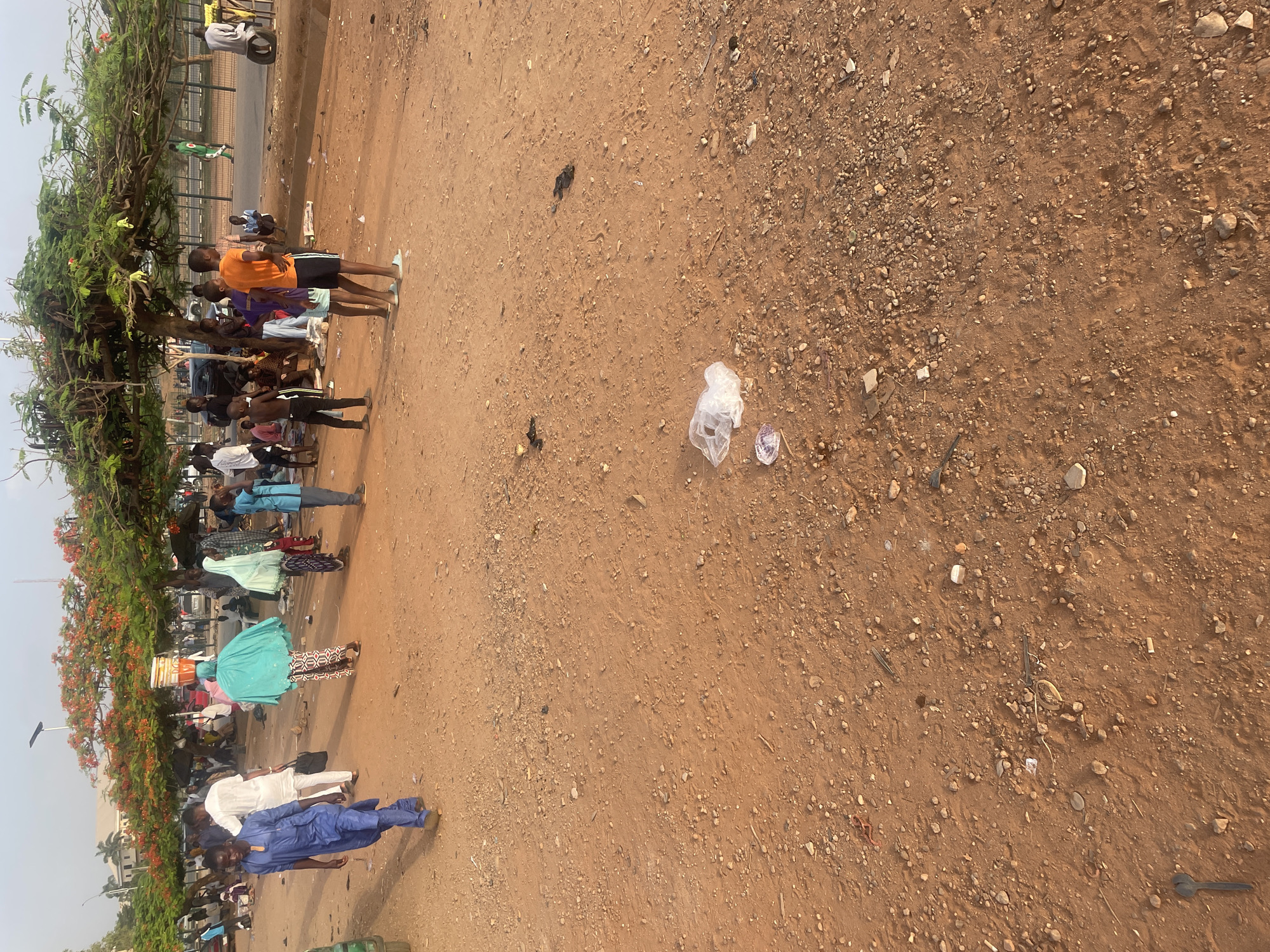 The image depicts a group of people congregated near a tree in an open, dusty area. The location appears to be a roadside or vacant lot in Abuja, Nigeria. The ground is primarily reddish-brown dirt and gravel, with scattered trash, including plastic bags and small debris. The people are diverse in age and dress, with some wearing casual attire and others in more traditional Nigerian clothing. Some individuals appear to be standing and observing, while others may be engaged in conversation or other activity. The background shows a road with vehicular traffic and possibly a fence or barrier. The overall scene suggests a typical gathering in a public space within an urban setting.