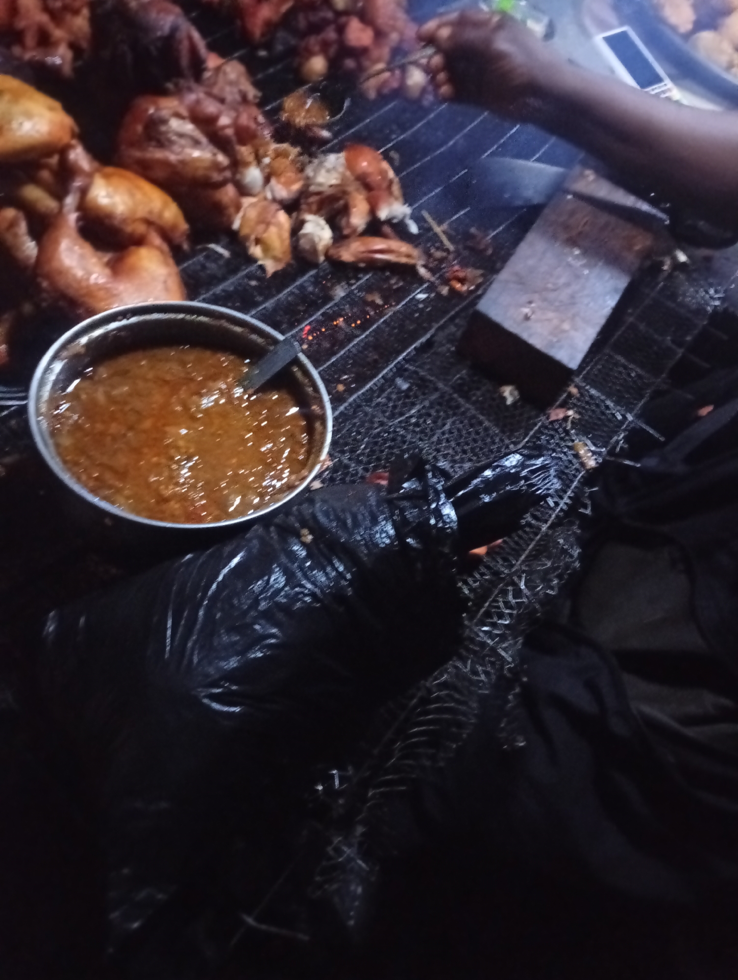 A close-up, dark, outdoor shot of food being prepared on a grill. In the foreground, a large plastic bag lies on a metal grate, obscuring part of the scene. To the left of the bag, a metallic bowl filled with a reddish-brown sauce sits on the grate. Behind the bowl, pieces of cooked chicken and other meats are visible on the grill. A person's arm and hand, holding a fork with food, are visible in the upper right quadrant. A wooden block is also on the grill. The overall impression is a busy food preparation area, likely at a street food stall. The location is not specified.