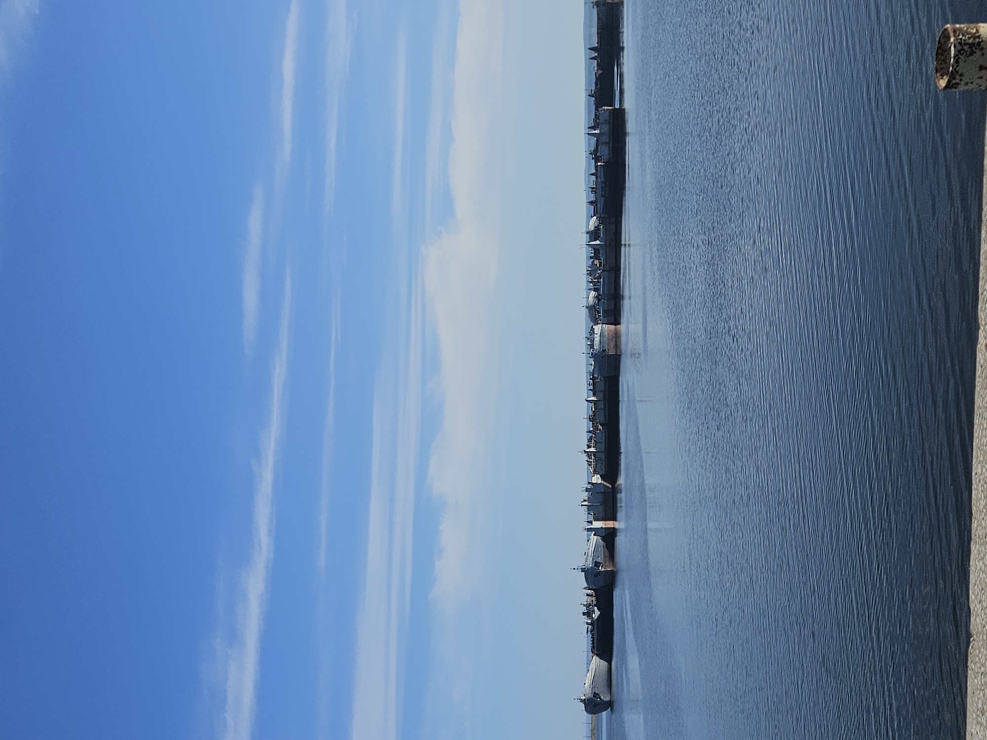 The image shows a view of the Kama River in Chaikovsky, Russia. The sky is a clear blue with some wispy clouds. The river is a darker blue and stretches out to the horizon. A line of ships and boats is anchored along the river. A rusty metal pole is visible in the right corner of the image. The picture appears to be taken from a high vantage point, possibly a bridge or tall building.