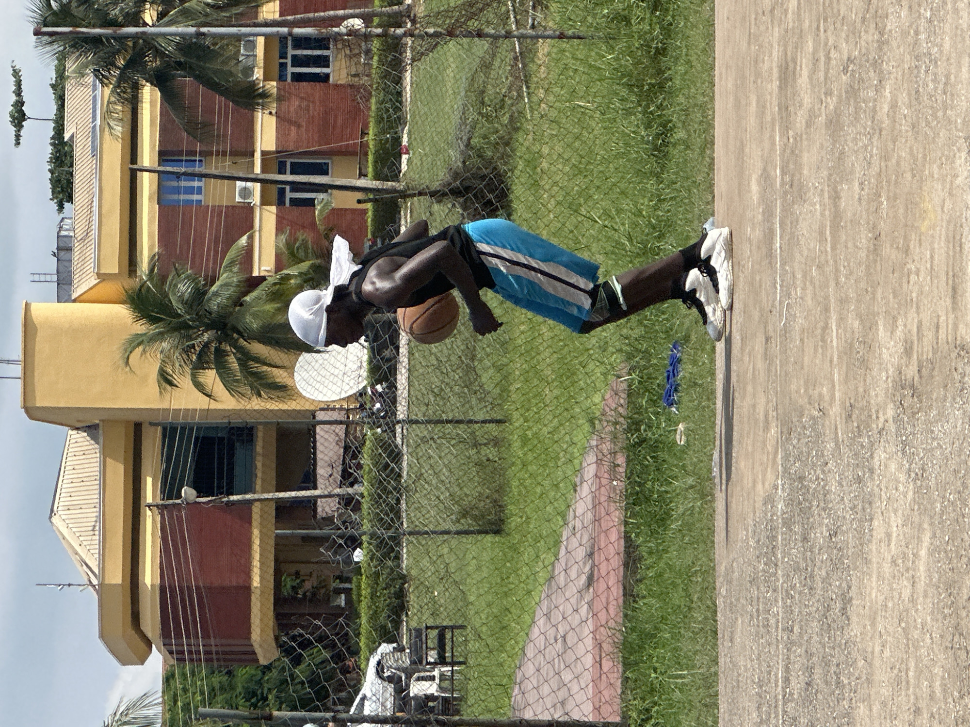 In Ajara, Nigeria, a young Black man with a white head covering dribbles a basketball on an outdoor court. He is wearing a black tank top and blue athletic shorts with white and black trim. He has black and white athletic shoes on his feet. The basketball court is made of concrete. Behind the court, a chain-link fence separates it from a grassy area. In the background, a large, yellow and red building with a light-colored roof stands under a cloudy sky. Palm trees are visible around the building. A white car and some motorcycles are parked in front of the building, and a white satellite dish is also visible.