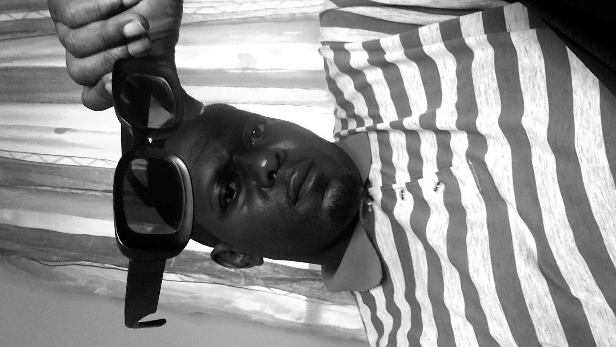 A grayscale portrait shows a man holding sunglasses. He is wearing a vertically striped shirt. The backdrop appears to be a wall or curtain with horizontal stripes. There is no information in the image to indicate the location is Sabon Tasha, Nigeria.