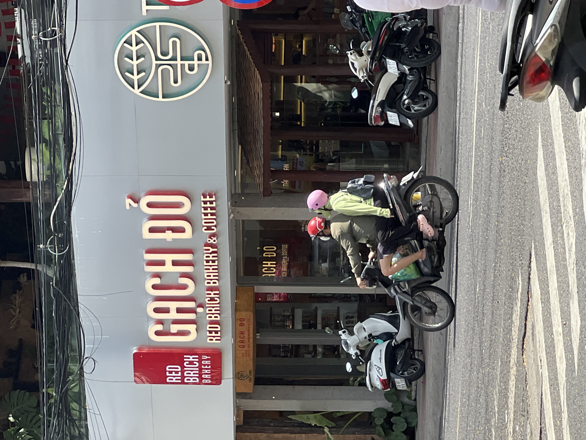 The image shows the exterior of a bakery and coffee shop in Nha Trang, Vietnam. The shop's name, "GACH DO RED BRICK BAKERY & COFFEE," is prominently displayed in large, illuminated letters on the facade. A logo is visible to the right of the name. Two individuals are riding a motorcycle in the foreground, passing in front of the shop. The rider is wearing a green jacket and a red helmet, while the passenger is wearing a light green top and a pink helmet. They are moving from left to right across the frame, with the motorcycle obscuring a portion of the shop's entrance. Several other motorcycles are parked along the sidewalk. The scene is set on a street with a crosswalk in the foreground. The time of day appears to be daytime, with natural lighting illuminating the scene.