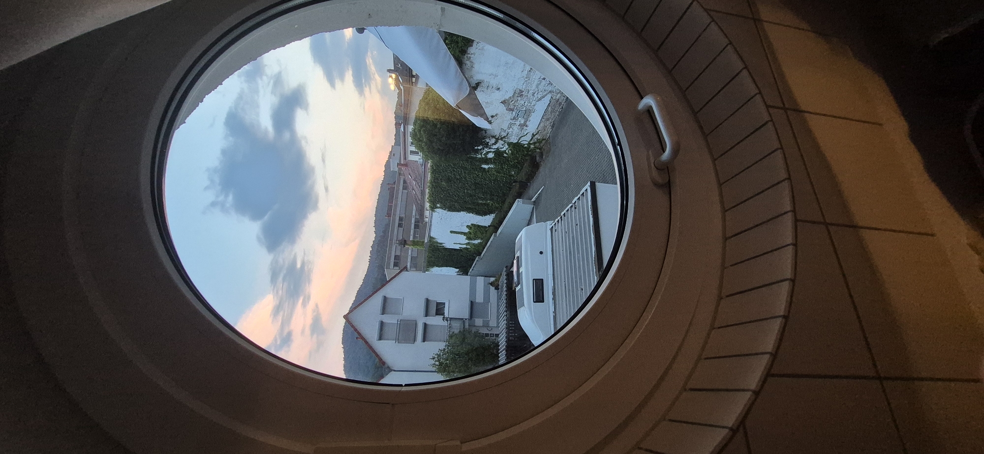 This image captures a view through a circular window looking out onto a residential area in Baden-Baden, Germany, during sunset. The foreground is dominated by the interior of the room, with a circular window frame and tiled sill.
Outside, the scene depicts a street or courtyard with a white house with grey shutters and a balcony visible. Adjacent to the house is a wall covered in dense green ivy, typical of European architecture. In the middle ground, a white trailer or camper van is parked. Beyond these elements, more buildings with red-tiled roofs are visible, along with a hill covered in trees in the background. The sky is painted with the warm hues of sunset, featuring soft pinks and oranges blended with blues and grey clouds. The lighting suggests it is late afternoon or early evening. No people are visible in the frame. The overall impression is of a quiet, residential neighborhood during a picturesque sunset.