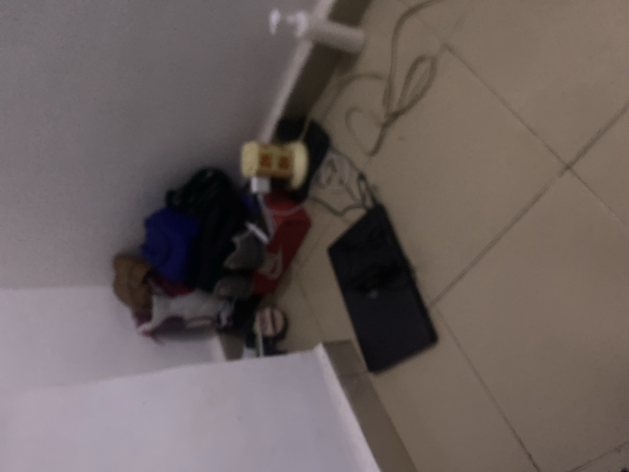 The image depicts a cluttered corner of a room with tiled flooring. In the background, against a white wall, several items are piled up, including what appear to be bags of clothing, some with a brown or tan hue, a dark blue garment, and a red box. There are also shoes visible, including a pair of grey sneakers and another pair of lighter colored sandals. To the right of this pile, on a black surface, is a cylindrical object with yellow and white markings, possibly a dispenser or a small appliance. Wires are spread across the floor in front of these items. In the foreground, a black laptop computer is placed flat on the floor. Further to the right, a white pump bottle is positioned against the wall. The overall scene suggests a disorganized living or storage space. The image is of low resolution and blurry, making precise identification of all objects difficult. The location of Howayi, Nigeria, is not visually discernible from the image content.