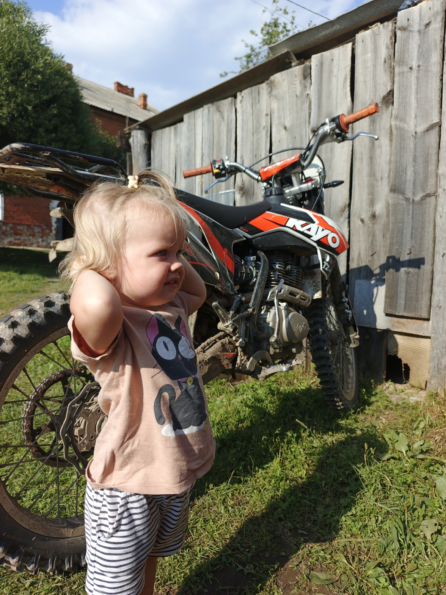 A young Caucasian female toddler with blonde hair pulled into a ponytail with a small yellow accessory stands in front of a dirt bike, leaning against a wooden fence. The toddler is wearing a light pink t-shirt with a black cat graphic and black and white striped shorts. Her arms are raised to her head, and she is looking to her right with an open mouth. The dirt bike is orange and black, with a Kayo logo visible on the side. The background consists of a wooden fence, green grass, and a cloudy blue sky. In the distance, a brick building with a grey roof and trees are visible. The scene appears to be outdoors in a rural setting, possibly a yard in Medvedyovka, Russia. The lighting suggests it is daytime, with shadows cast on the ground.