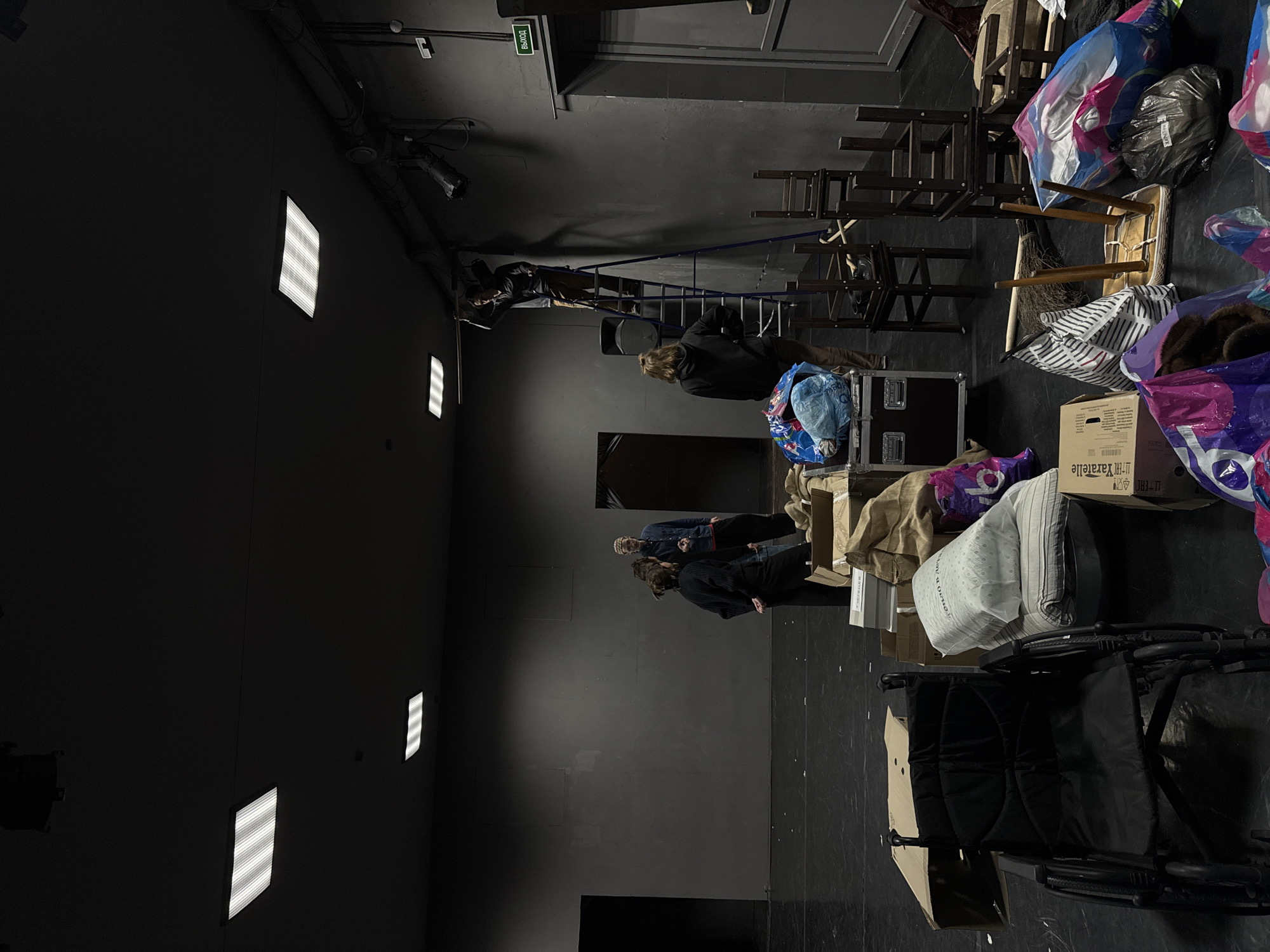 The image captures an indoor scene, likely a storage or backstage area within a theater or event venue, characterized by dark gray or black walls and floor. The space is illuminated by overhead stage lights and several rectangular fluorescent light fixtures mounted on the left wall, casting a dim, utilitarian ambiance.
Several individuals are present, seemingly engaged in organizing, moving, or preparing items. On the left, one person is standing on a blue ladder, possibly attending to overhead equipment. Further to the right, other individuals are standing amidst various objects, their faces mostly obscured.
The room is cluttered with numerous items. Stacks of dark wooden chairs are prominent in the center-right. There are also multiple cardboard boxes, some open, some closed, and numerous plastic and fabric bags, many appearing full, suggesting they contain clothes or other soft goods. A black equipment case and a dark stroller or child carrier are also visible on the right side of the frame. A broom leans against some stacked chairs.
A green exit sign with the word "ВЫХОД" (Russian for "EXIT") is clearly visible on the top left, indicating a Russian-speaking location, aligning with the provided context of Moscow, Russia. On one of the cardboard boxes, partially obscured text reads "ТТ-ЕЙС Хаratejle". The scene depicts a functional, logistical activity, possibly related to an event setup, teardown, or general storage management. No specific emotional expressions are discernible among the individuals.
