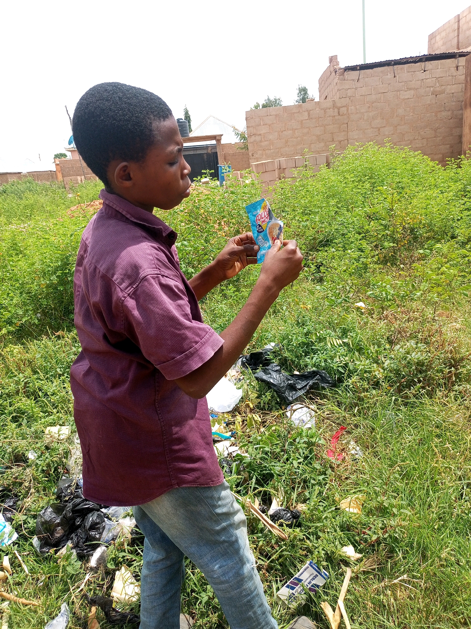 A young boy of African descent, wearing a maroon short-sleeved collared shirt and blue jeans, stands in a grassy area littered with debris. He is holding a small packet of "GOLDEN MOMENT" snack in his hands. In the background, there are unfinished brick buildings and scattered vegetation. The scene appears to be an outdoor location in Tempure, Nigeria.