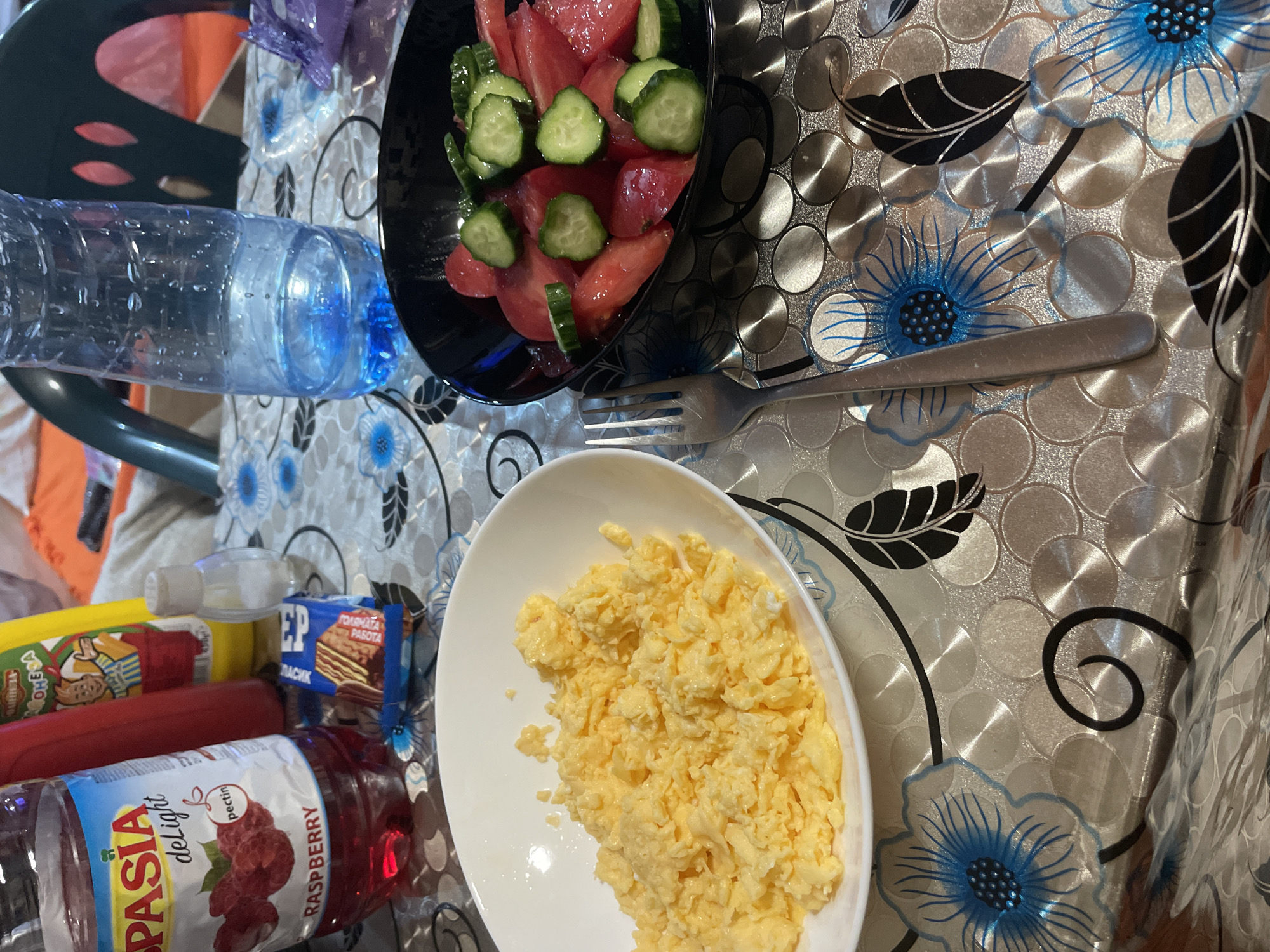 The image displays a table set with food and beverages, suggesting a meal in an indoor setting. The table is covered with a reflective, patterned tablecloth featuring silver circular motifs and blue floral designs with black leaves.
In the center, a white oval plate holds a serving of fluffy scrambled eggs. To its left, a clear plastic bottle containing a red liquid, labeled "SPASIA delight" with "pectin" and "RASPBERRY" printed on it, lies horizontally. Adjacent to this, a small blue packet of wafers or biscuits is visible, with Cyrillic text reading "ЗЕР" (ZER), "ГОЛЕМАТА РАБОТА" (GOLEMATA RABOTA - "The Big Work/Thing"), and "КЛАСИК" (KLASIK - "Classic"). A yellow squeeze bottle, possibly containing a condiment, shows partially visible Cyrillic text "ОМЕГА" (OMEGA).
Further back, a black bowl contains a salad composed of diced red tomatoes and sliced green cucumbers. A silver fork rests between this bowl and the scrambled eggs. To the left of the salad, a clear plastic bottle of water is partially visible. The background includes glimpses of an orange object, possibly a chair or fabric, and an indistinct indoor environment. The ambient lighting suggests it is daytime. No people are present. The Cyrillic text on the packaging aligns with the given location context of Ihtiman, Bulgaria.
