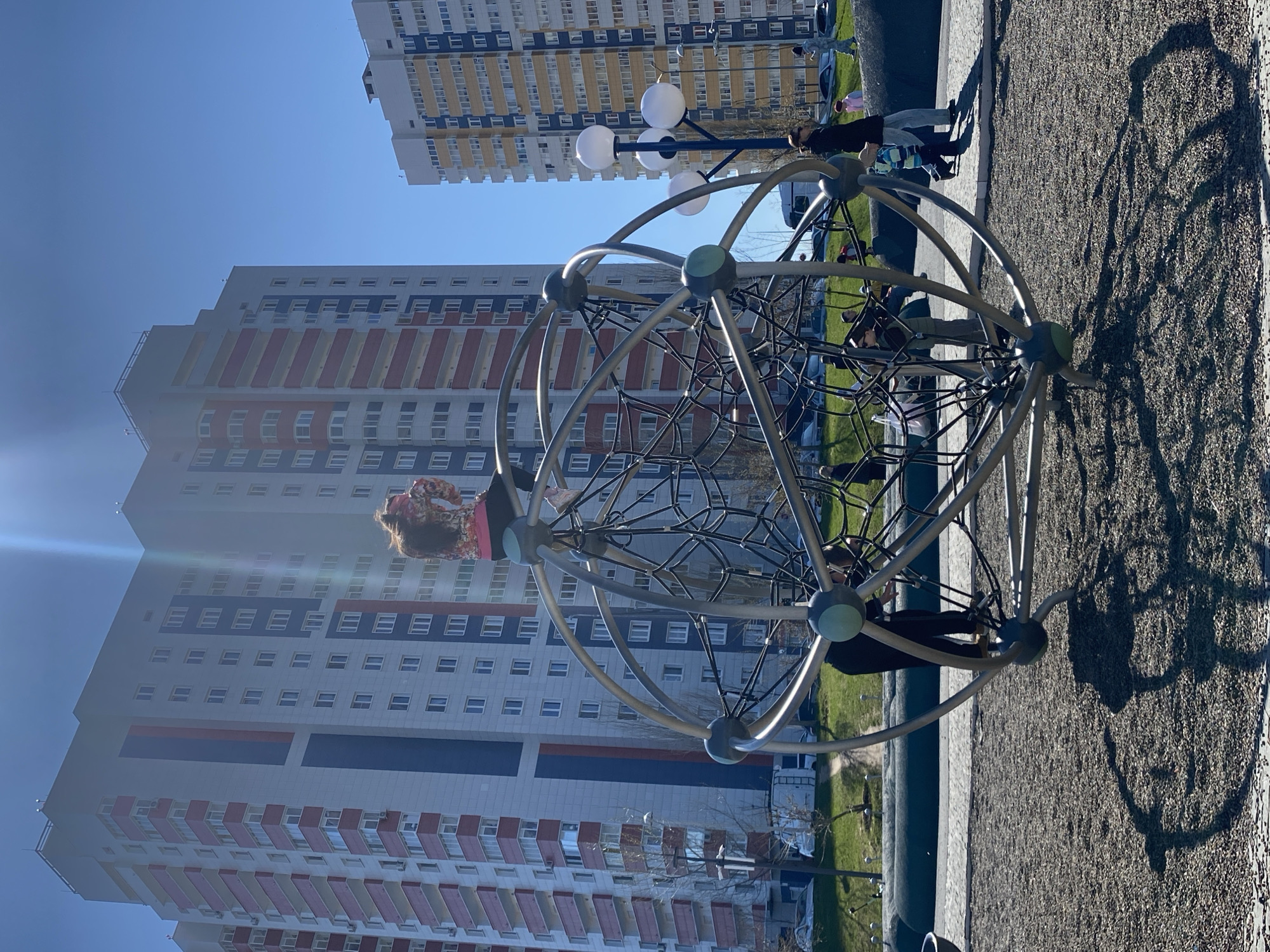 The image shows a playground in the city of Kazan, Russia, located outdoors. In the foreground is a spherical climbing frame made of metal bars and ropes. A young girl wearing a floral shirt and black pants is climbing on the frame, with another person visible behind her. The playground surface appears to be covered with gravel.
In the background, there are several tall apartment buildings with multiple floors and numerous windows. A row of streetlights is positioned to the side, adding to the urban setting. The sky above is clear and blue. The scene appears to be shot during the daytime, with the sun casting shadows of the climbing frame on the playground surface.