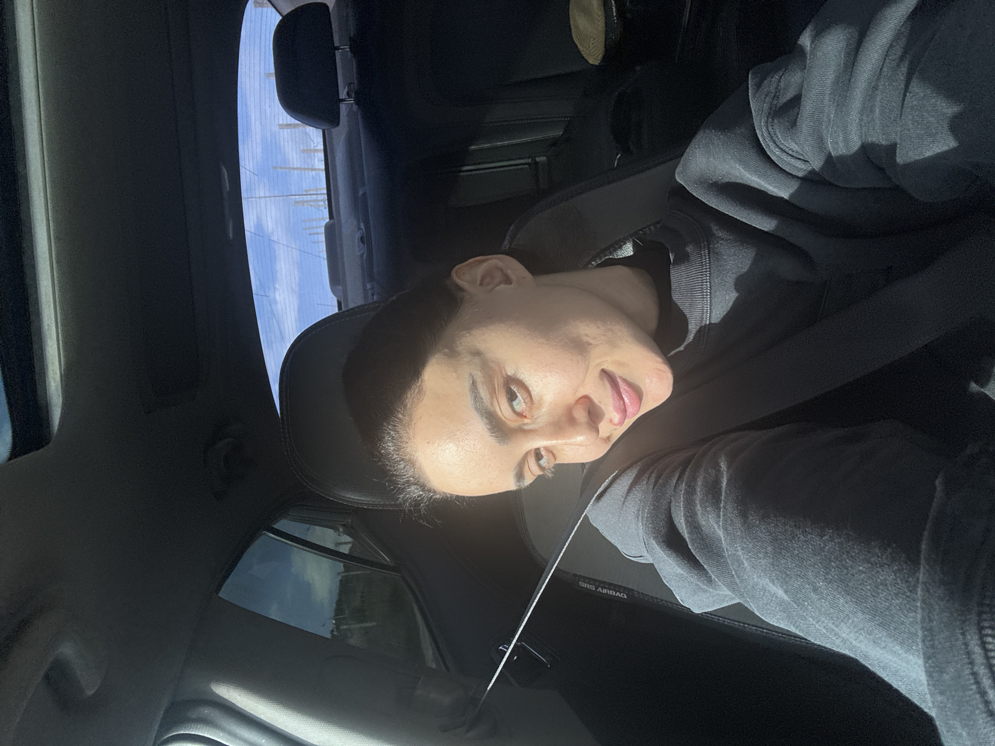 A person is sitting inside a car, wearing a seatbelt and a dark hooded sweatshirt. The car's interior is dark, and sunlight is streaming through the windows. Outside the car windows, tall poles can be seen against a light blue sky, suggesting a location near infrastructure like power lines or a port. Due to the visible infrastructure, the location is potentially in Tbilisi, Georgia.
