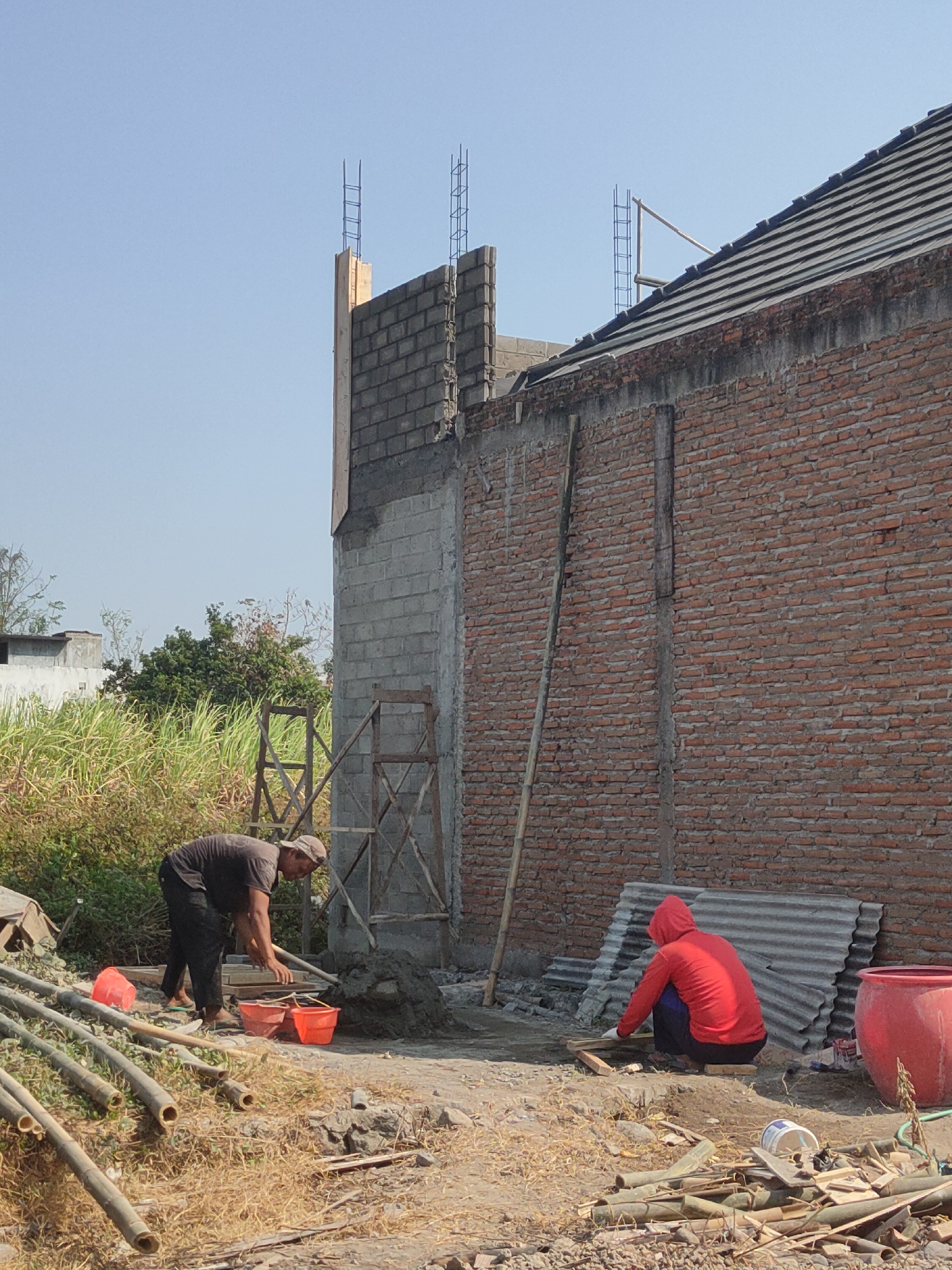 The image depicts an outdoor construction site in Ngasem, Indonesia. Two male construction workers are actively engaged in their tasks. One worker, wearing a dark t-shirt and cap, is bent over, appearing to mix or handle construction materials near two orange buckets and a red bucket. The second worker, dressed in a red hooded jacket and dark pants, is crouched down, working on something with wooden planks in front of a corrugated metal sheet.
The environment is characterized by unfinished brick and concrete structures. A red brick wall forms a prominent feature of a building under construction. Rebar is visible extending from the top of a concrete pillar and the edge of the building. Other objects scattered around the site include bamboo poles, construction debris, and a large red barrel. The ground is a mixture of dry earth and grass. The sky is clear and blue, indicating daytime. Lush green vegetation, including tall grass, is visible in the background, suggesting a rural or semi-rural setting.