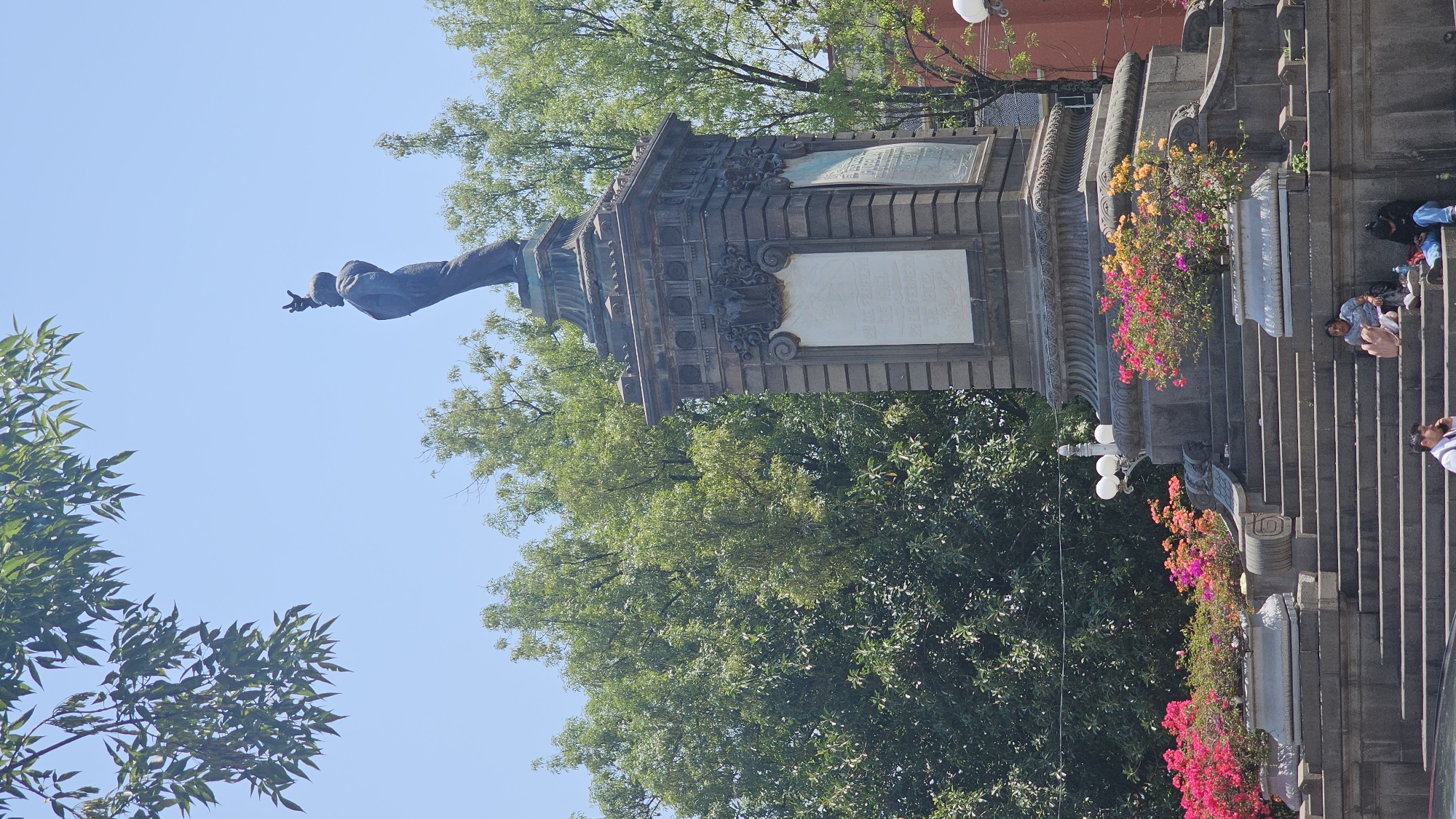The image displays a prominent historical monument under a clear blue sky, likely situated in a public plaza in Puebla, Mexico, as suggested by the context. The monument consists of a multi-tiered, intricately designed stone base adorned with ornate carvings and several rectangular plaques. These plaques, though illegible, appear to bear inscriptions or dedications.
Crowning the monument's base is a bronze statue of a male figure, depicted in formal attire with one arm outstretched and hand gesturing, indicative of an orator or a commanding presence. Surrounding the monument and extending into the background are lush green trees and foliage, with some branches framing the blue sky.
Stone steps leading up to the monument's base are visible on the right side of the image. Several individuals are seated on these steps, appearing relaxed and enjoying the sunny weather. The steps are decorated with planters filled with vibrant flowers in shades of pink, orange, and yellow, adding splashes of color to the stone architecture. Partial views of streetlights with white globes are also visible near the steps.
The scene is bathed in bright daylight, suggesting it is midday or afternoon, with clear weather. The overall impression is one of a peaceful public space, where history and natural beauty converge, and people gather to relax. There is no clearly readable text on the plaques due to the distance and angle.