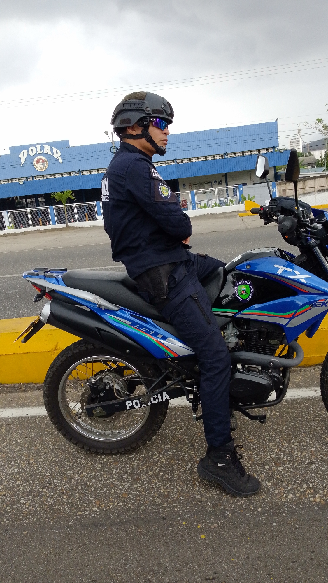 A uniformed police officer is seated on a blue and black motorcycle along a road. The motorcycle is labeled "Policia" on the rear. The officer wears a dark blue uniform, black boots, a tactical helmet, and sunglasses. A pistol holster is visible on their right hip. Behind the officer and motorcycle, a building with "Polar" signage is visible. The photo was taken in the city of Tinaquillo, Venezuela.