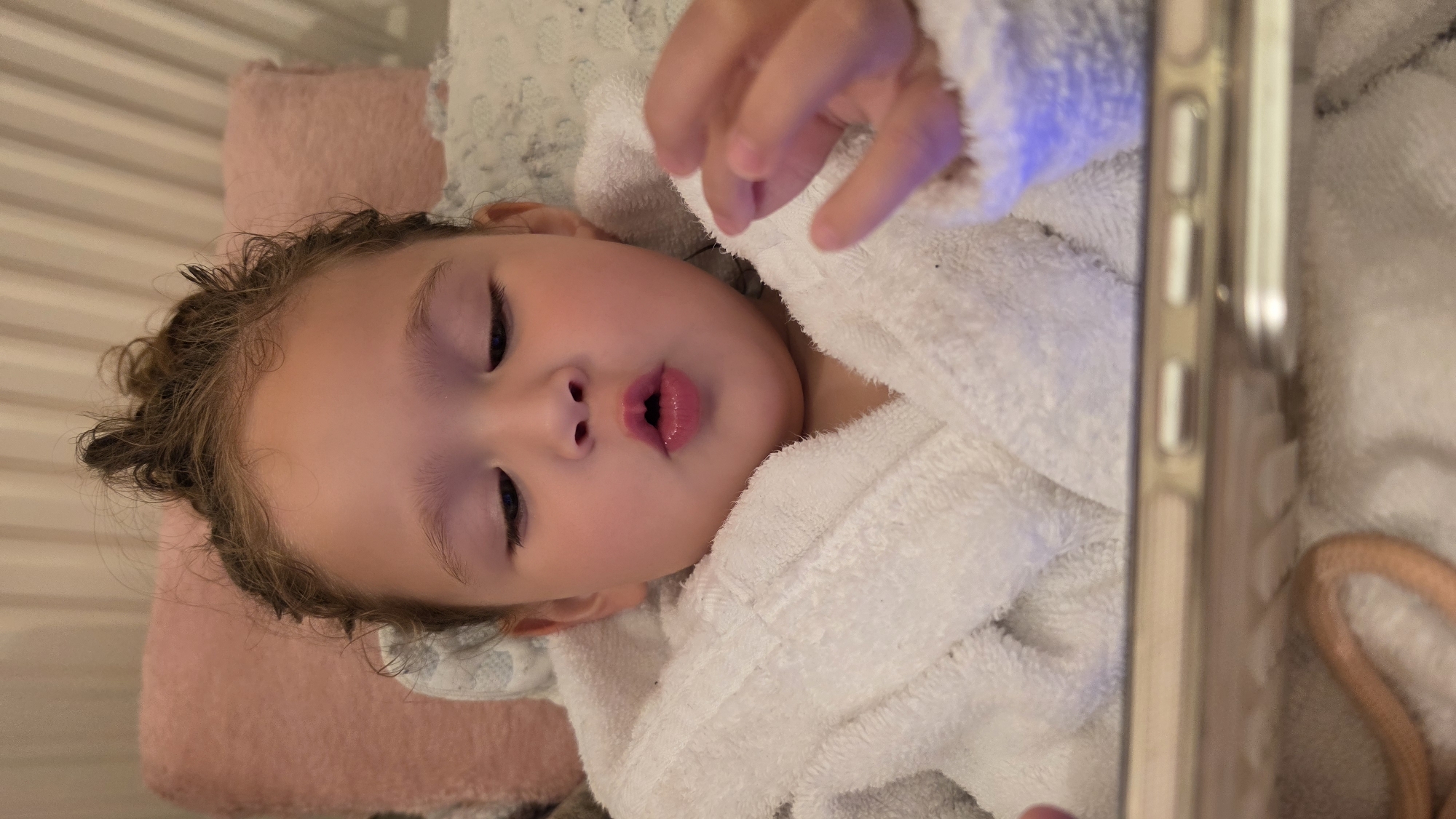 A young child with curly brown hair, wearing a white bathrobe, lies on a pink pillow with a gray and white patterned blanket partially underneath the pillow. The child's eyes are closed, and the lips are slightly parted. Part of a mobile phone is visible on the right side of the frame. The setting appears to be indoors, possibly a bedroom.
There is no information to suggest that the location is in the city of London, United Kingdom.