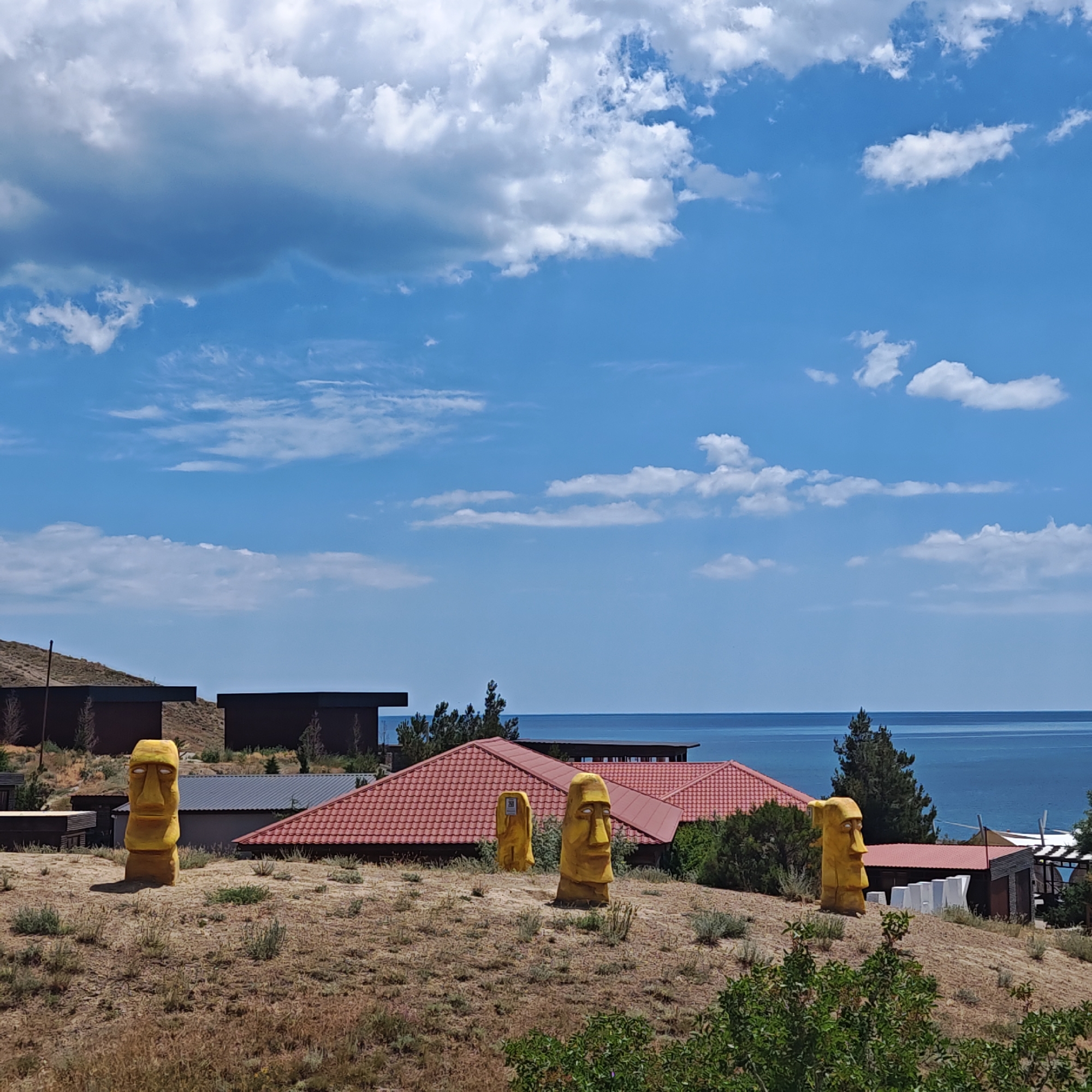 The image depicts an outdoor scene with a view of the sea in the background. In the foreground, there are several yellow, Moai-like statues placed on a dry, grassy hillside. Behind the statues, there are buildings with red tiled roofs and dark colored structures. The sky is blue with scattered white clouds. The location is identified as Sudak, Ukraine. There are no people visible in the image. The environment appears to be somewhat arid, with dry vegetation and sparse bushes. The sea is calm and stretches towards the horizon. The overall scene suggests a unique tourist attraction or a stylized residential area near the coast.