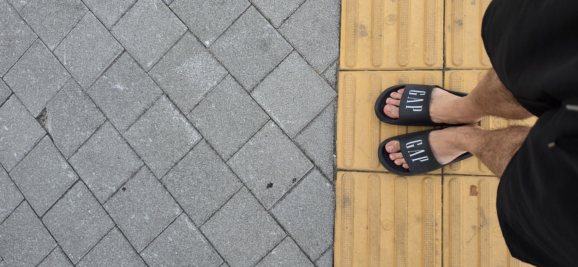 A first-person perspective shot shows a person wearing black GAP slides standing on a textured yellow tactile paving sidewalk adjacent to a gray stone-tiled pavement. The person is wearing black shorts. The scene appears to be outdoors in an urban setting, likely on a sidewalk in Cheongju-si, South Korea, given the typical design of tactile paving used for the visually impaired. The lighting suggests it is daytime.