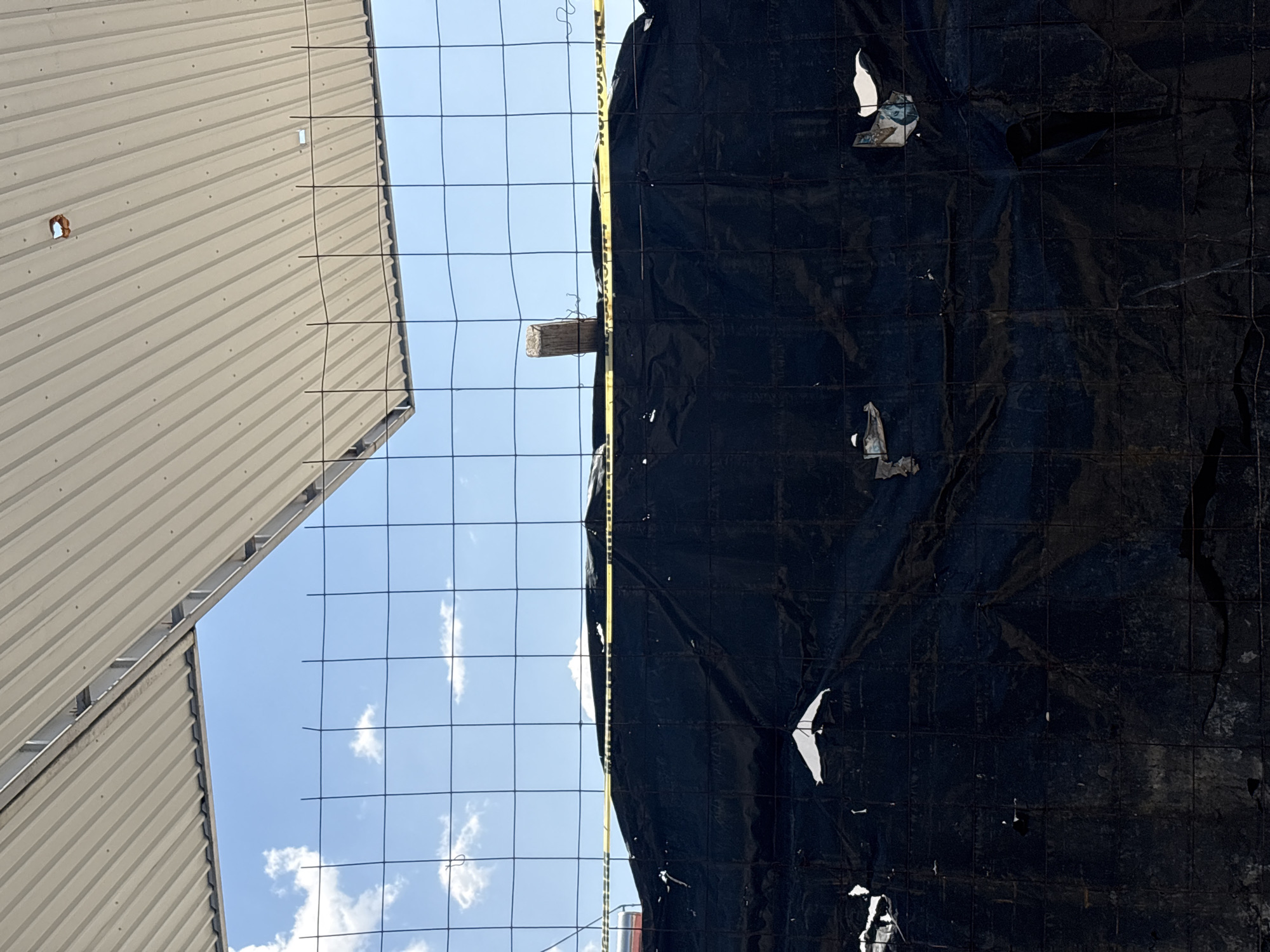 The image displays an outdoor construction or restricted area in Puebla, Mexico, under a clear blue sky with scattered white clouds. The upper frame shows a section of a light-colored corrugated metal roof structure with an open corner revealing the sky. A small, irregular brown object is affixed to the roof. A black square-grid wire mesh fence horizontally spans the mid-ground. Behind the fence, a dark, heavily wrinkled black tarpaulin serves as a barrier, displaying multiple tears that expose lighter material beneath. A yellow caution tape with black Spanish lettering is stretched horizontally across the top of the tarp, visible through the fence. A single, rough wooden post protrudes vertically from behind the tarp on the right side. No individuals or specific actions are visible.