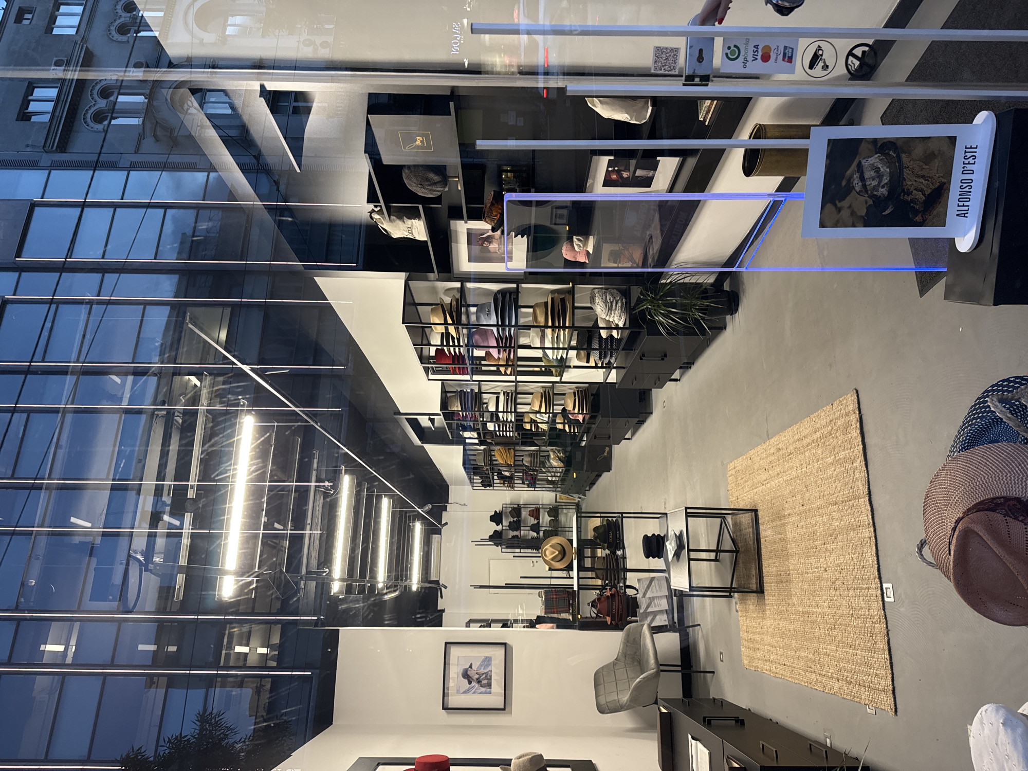 The image captures the interior of the ALFONSO D'ESTE hat store in Belgrade, Serbia, as seen through the storefront window. The store features shelves displaying various hats, interspersed with decorative items and framed photographs. A rug lies on the floor near a small table. The reflection in the window reveals the exterior architecture of a building with multiple windows and details, contrasting with the modern interior of the shop. No people are directly visible in the scene.