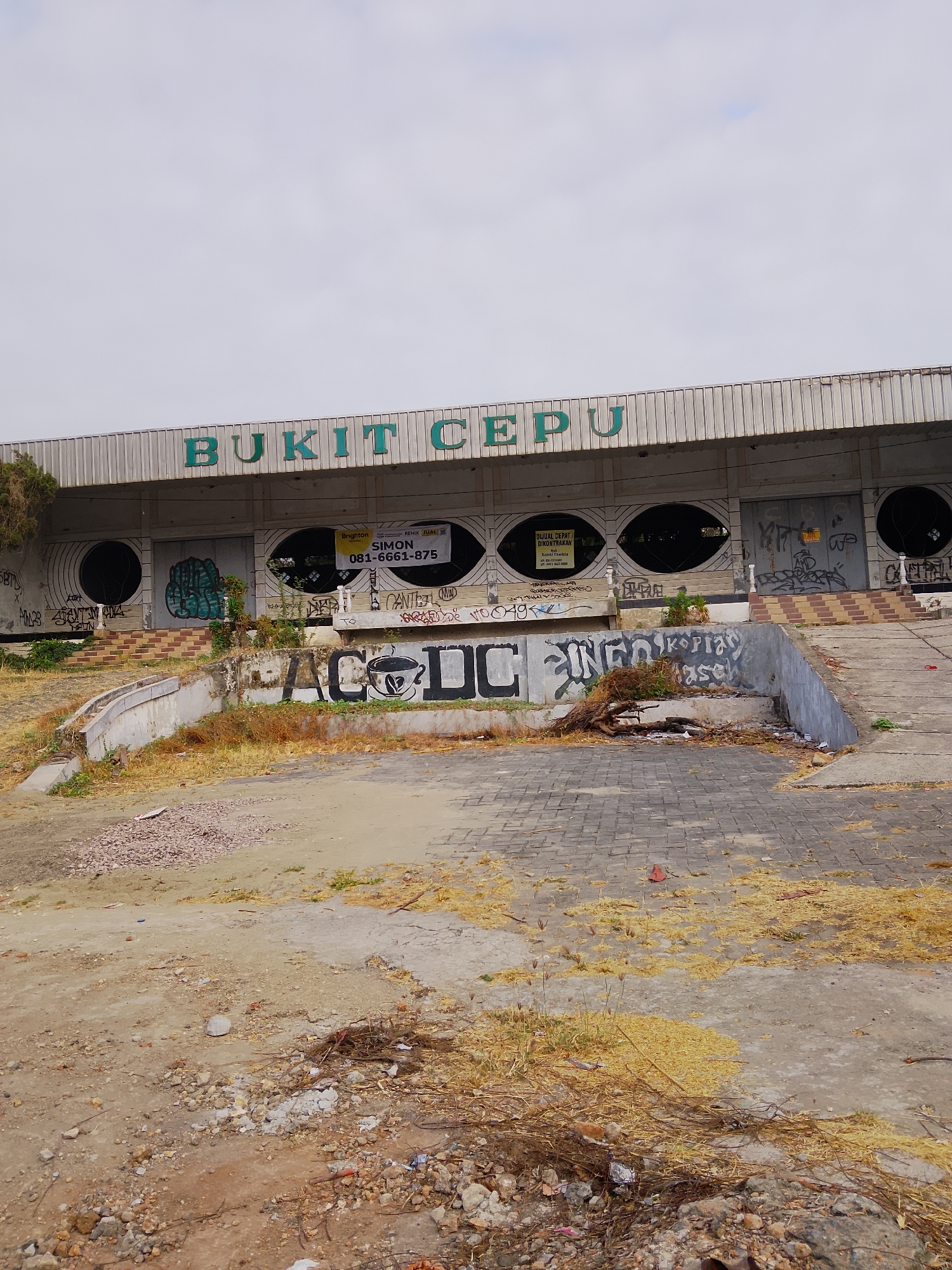 The image depicts an abandoned building with the words "BUKIT CEPU" prominently displayed in green letters above a row of large, oval-shaped openings. The building's facade is covered in graffiti, with various tags and drawings visible. A yellow "DIJUAL/DIKONTRAKAN" (For Sale/For Rent) sign is affixed to the building, along with a smaller sign listing a phone number. The ground in front of the building is unkempt, with dry grass, debris, and rubble scattered across a paved area and dirt patches. Steps leading up to the building are also visible, some with brick and concrete elements. The overall scene suggests a state of neglect and disrepair in what appears to be a semi-urban or commercial area.