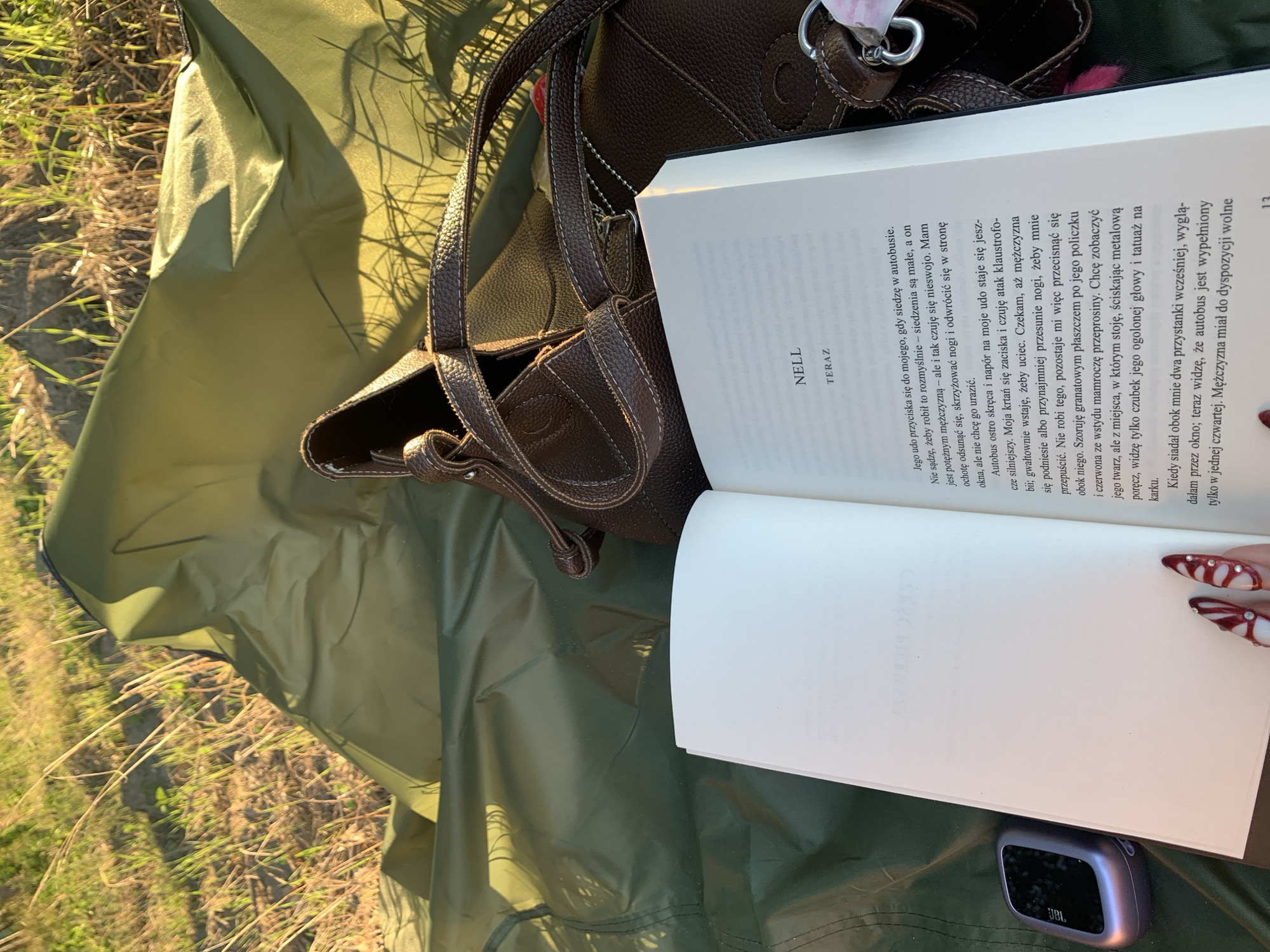This image depicts an outdoor scene during daytime, with a focus on a book, a brown leather handbag, and a small electronic device laid out on a green fabric. The book is open to a page with text, and the title "NELL" is visible, followed by "TERAZ". The surrounding environment appears to be a grassy area with dry reeds and sunlight casting shadows, suggesting a natural setting. There is no indication of the city of Massenzatica, Italy, or any specific urban location. The image is captured from a slightly elevated perspective, looking down onto the arrangement of objects. No people are directly visible in the frame, although a fingernail with nail art is partially visible at the bottom of the frame, suggesting a human presence. The overall mood is relaxed and suggests a moment of leisure.