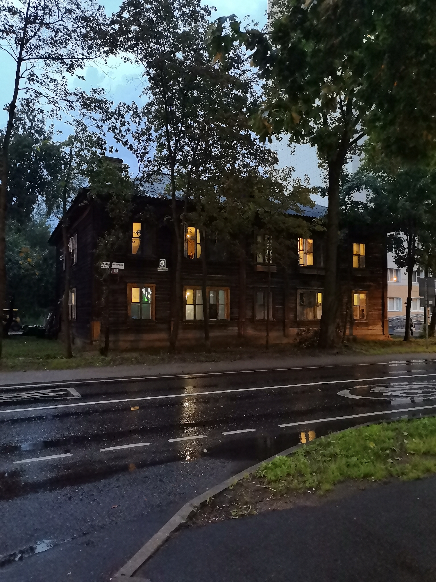 The image captures a scene on a wet street in Krasnogorsk, Russia, likely during twilight or overcast conditions. A two-story wooden building, appearing aged and dark, stands prominently on the left side of the frame. Its windows are illuminated from within, casting a warm glow. Trees with dense foliage surround the building and line the street. A wet asphalt road with white lane markings and a circular speed limit sign reflects the dim light and scattered puddles. In the background, a modern multi-story building with illuminated windows suggests a contrast between older and newer urban development. No individuals are clearly visible in the scene.
