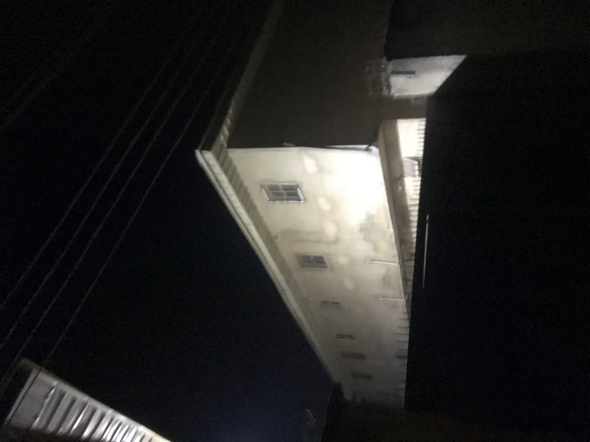 The image depicts the exterior of a light-colored building at night. The building appears to be multi-storied, with several rectangular windows visible along its facade. The windows are dark, suggesting they are either closed or the interior is not illuminated. Power lines are visible overhead, crisscrossing the dark night sky. In the foreground, a dark, solid object, possibly a wall or fence, obstructs the lower portion of the view. The overall scene is dimly lit, with artificial light sources implied but not directly visible, casting a pale glow on the building's surface. The location is identified as Rafin Par, Nigeria.