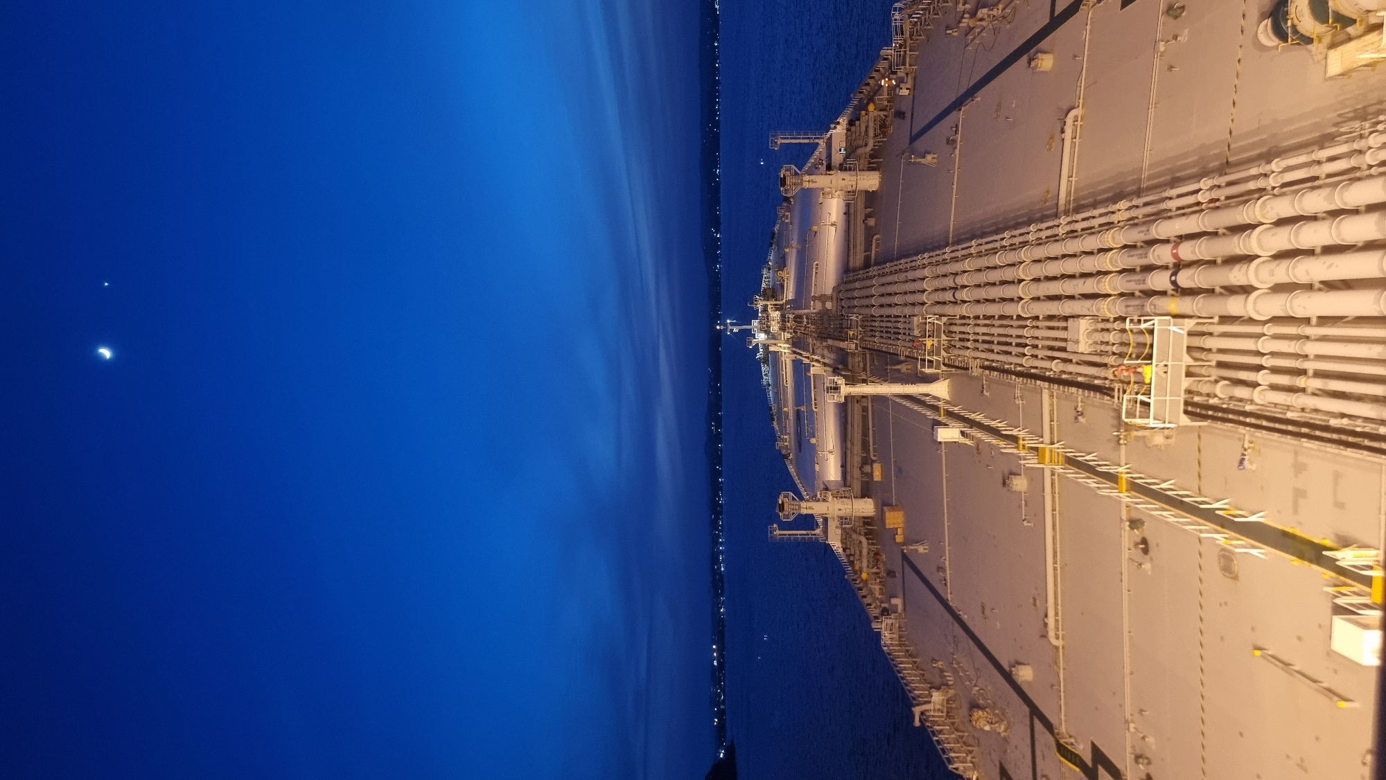 The image shows a large oil tanker at sea at dusk. The sky is a deep blue, with a crescent moon and a bright star visible. The sea is calm, and in the distance, the lights of a city can be seen along the coastline. The deck of the tanker is covered in a complex network of pipes and equipment. A small platform with two figures in safety vests is visible on the right side of the deck, near the pipes. The overall scene suggests a peaceful and industrial setting at the end of the day. The image is consistent with a view from the bridge of a vessel in the port of Trincomalee, Sri Lanka, given the presence of a large port and a natural harbor that accommodates large vessels like oil tankers.