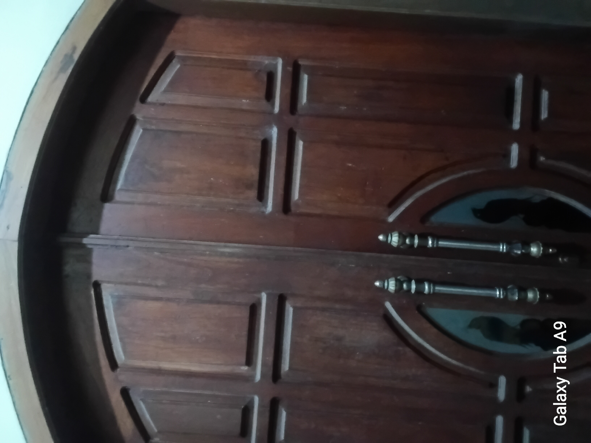 The image shows a close-up view of a large, wooden double door with a curved top. The doors are dark brown and have recessed panels with horizontal rectangular indentations. Two decorative door handles, made of metal with a brass finish, are visible in the lower half of the image. The handles are ornate, with bulbous decorative elements. An oval-shaped glass insert is present on each door, behind the handles, and appears to be tinted or reflective. The overall impression is of a substantial and possibly traditional entryway. There is a watermark in the lower-left corner that reads "Galaxy Tab A9". The location is identified as Trenggalek, Indonesia.