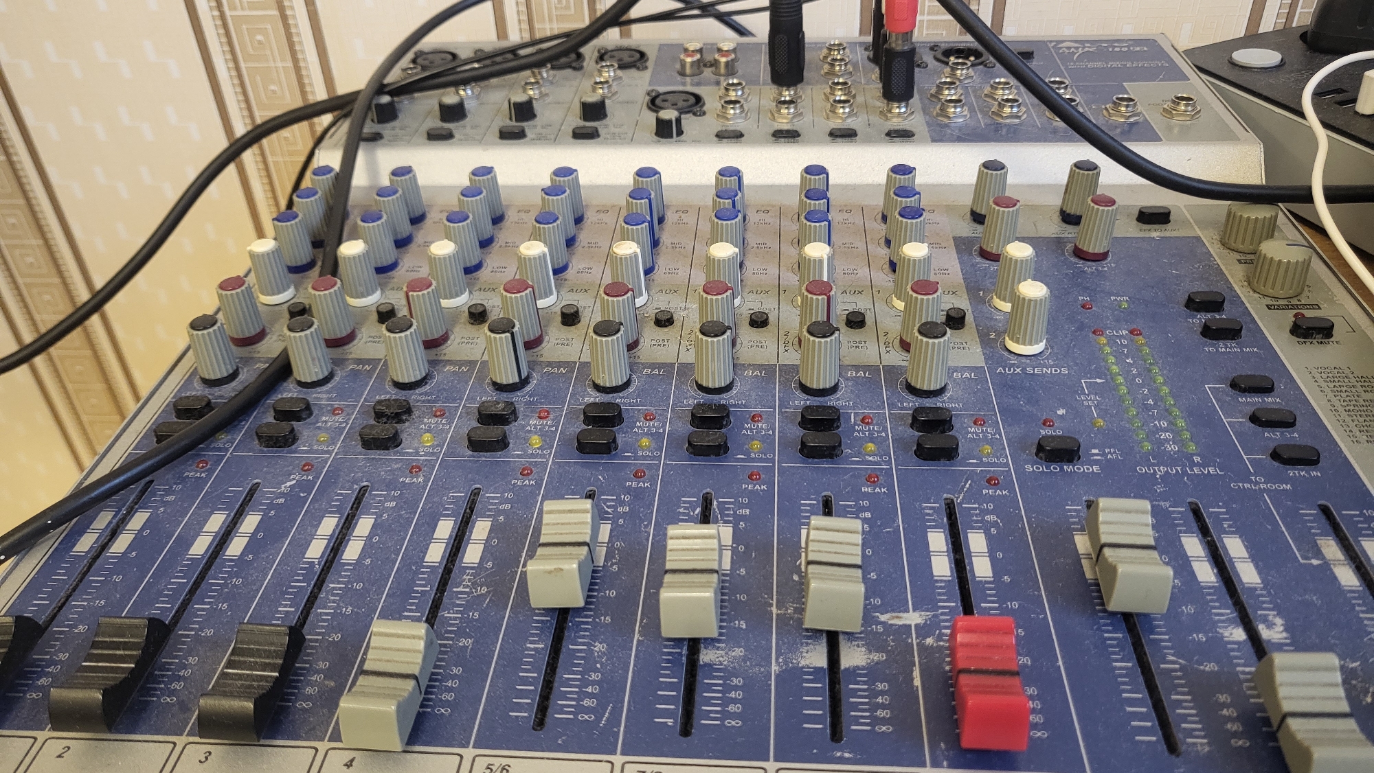 The image captures a detailed, eye-level view of an audio mixing console, likely in an indoor setting such as a studio or control room in Марковский, Russia. The main subject is a blue and gray multi-channel mixer, densely populated with controls.
The console features multiple vertical faders, predominantly light gray with one notable red fader on the right. These faders operate along calibrated tracks marked from -60 dB to +10 dB, each with a "PEAK" indicator. Above the faders are rows of black push-buttons labeled "MUTE/ALT 3-4" and "SOLO." Further up, numerous rotary knobs, some with blue, white, or red caps, control functions such as "EQ" (with frequency labels like "LOW 80Hz" and "MID 2.5KHz"), "AUX," "PAN," and "BAL" (Balance). Channels are numbered, with "2," "3," "4," and "5/6" visible.
In the central right section, a vertical LED meter displays "OUTPUT LEVEL," ranging from -30 to +10, with a "CLIP" warning light. This area also includes labels for "AUX SENDS," "LEVEL SET," "SOLO MODE," "MAIN MIX," "ALT 3-4," and "TO CTRL-ROOM." A larger gray knob labeled "VARIATIONS" and a "DFX MUTE" button are present, alongside a partially visible list of digital effects such as "VOCAL 1," "LARGE HALL," "PLATE," and "REVERS."
Behind the primary mixer, a smaller, lighter-colored "ANALOG MIXING CONSOLE WITH DIGITAL EFFECTS" is partially visible. Several black and one white audio cables connect the devices. The equipment appears used, with visible dust on the surfaces, suggesting a working environment. The background shows patterned wallpaper, indicating a functional rather than pristine setup. No individuals are present, but the arrangement suggests the equipment is set up for audio mixing, recording, or live sound production.