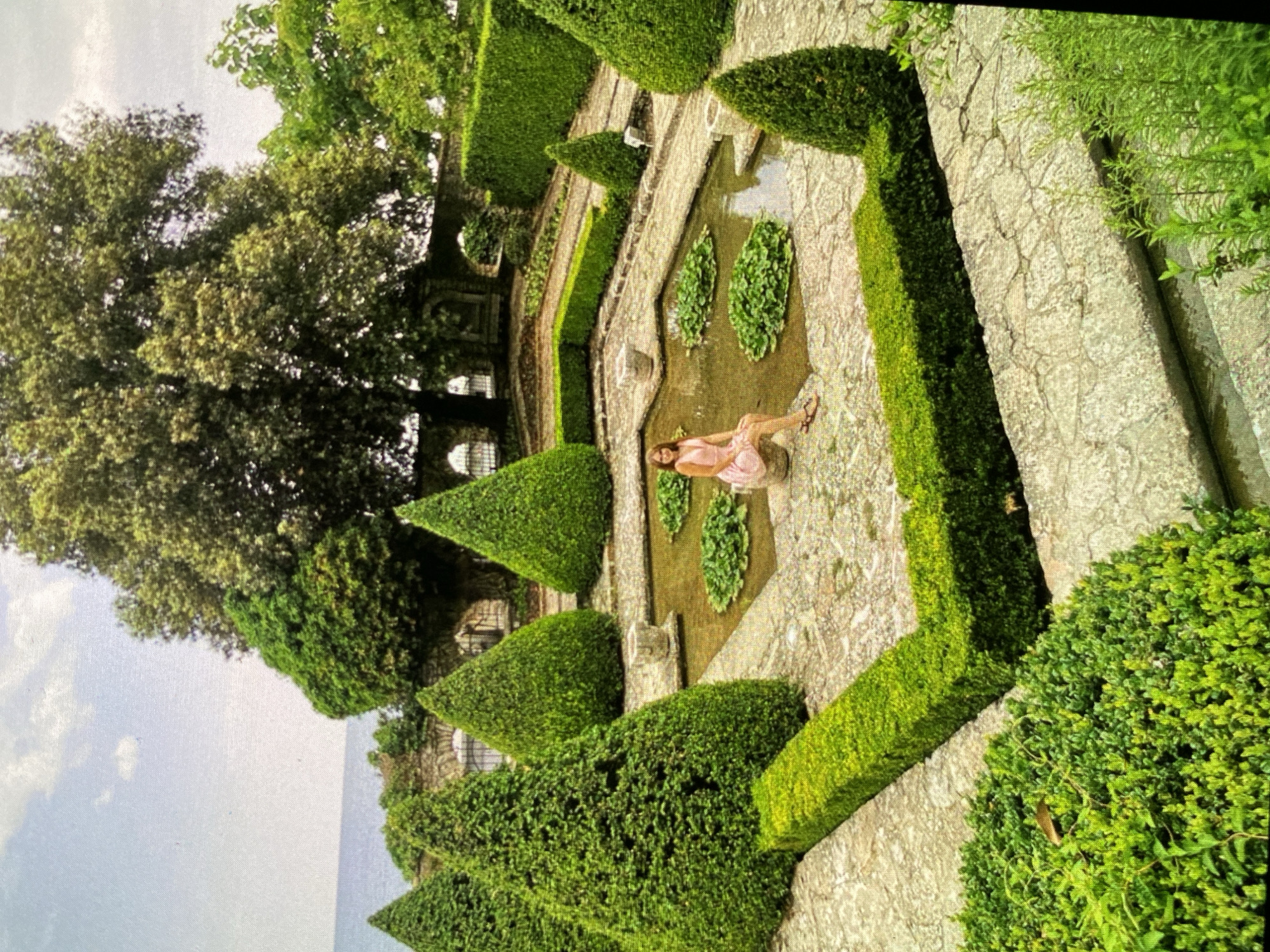 This image, rotated counter-clockwise, captures a serene formal garden featuring meticulously sculpted topiary hedges and stone pathways. A woman in a light-colored (possibly pink or peach) dress is gracefully seated on the edge of a rectangular water feature, which contains several aquatic plants resembling lily pads. She appears relaxed, with her legs crossed and wearing sandals.
The garden showcases a classic design with intricate patterns of conical and rectangular hedges of varying heights, framing the central water element. Large, mature trees with lush green canopies are prominent in the upper left background. In the distance, beyond the garden's edge, a broad expanse of blue is visible, indicative of a large body of water, such as a lake or sea, beneath a partly cloudy sky. Stone walls and architectural elements are partially visible amidst the foliage. The scene is illuminated by bright daylight, suggesting late morning or afternoon, with soft, ambient lighting. The overall atmosphere conveys tranquility and the beauty of a well-maintained landscape. No text is visible in the image.