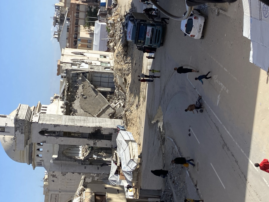 The image depicts an urban street scene in Khan Yunis, Palestinian Territory, characterized by widespread destruction under clear skies. A large, multi-story building, identified as the Al-Farouq Mosque due to its distinctive dome and minaret structure, occupies the left and central background. This building exhibits severe structural damage, with significant portions of its facade and interior walls collapsed, leaving exposed concrete and twisted rebar. Piles of rubble and debris surround the base of the damaged building.
In the midground, several makeshift shelters constructed from white tarpaulins are erected on sandy, uneven ground amidst debris, positioned near the damaged mosque.
A paved asphalt road, with faded white lane markings and visible wet patches or puddles, curves through the foreground and midground. On the road, multiple individuals are present. In the immediate foreground, two children are playing: one child in a brown top is on the ground near a small, dark object (possibly a ball), and another child in a blue top is running nearby. A third child in a red top is partially visible in the bottom left. An adult male carrying a yellow bag walks on the left side of the road, and a woman in a black abaya walks further back.
On the right side of the road, a blue and white semi-truck is either parked or moving slowly, with several people gathered around it or walking past. A white compact car is parked on the far right. In the background, beyond the immediate destruction, other urban buildings, some appearing intact and others potentially damaged, are visible under the bright sunlight. Various individuals are scattered throughout the scene, observing, walking, or engaging in activities.