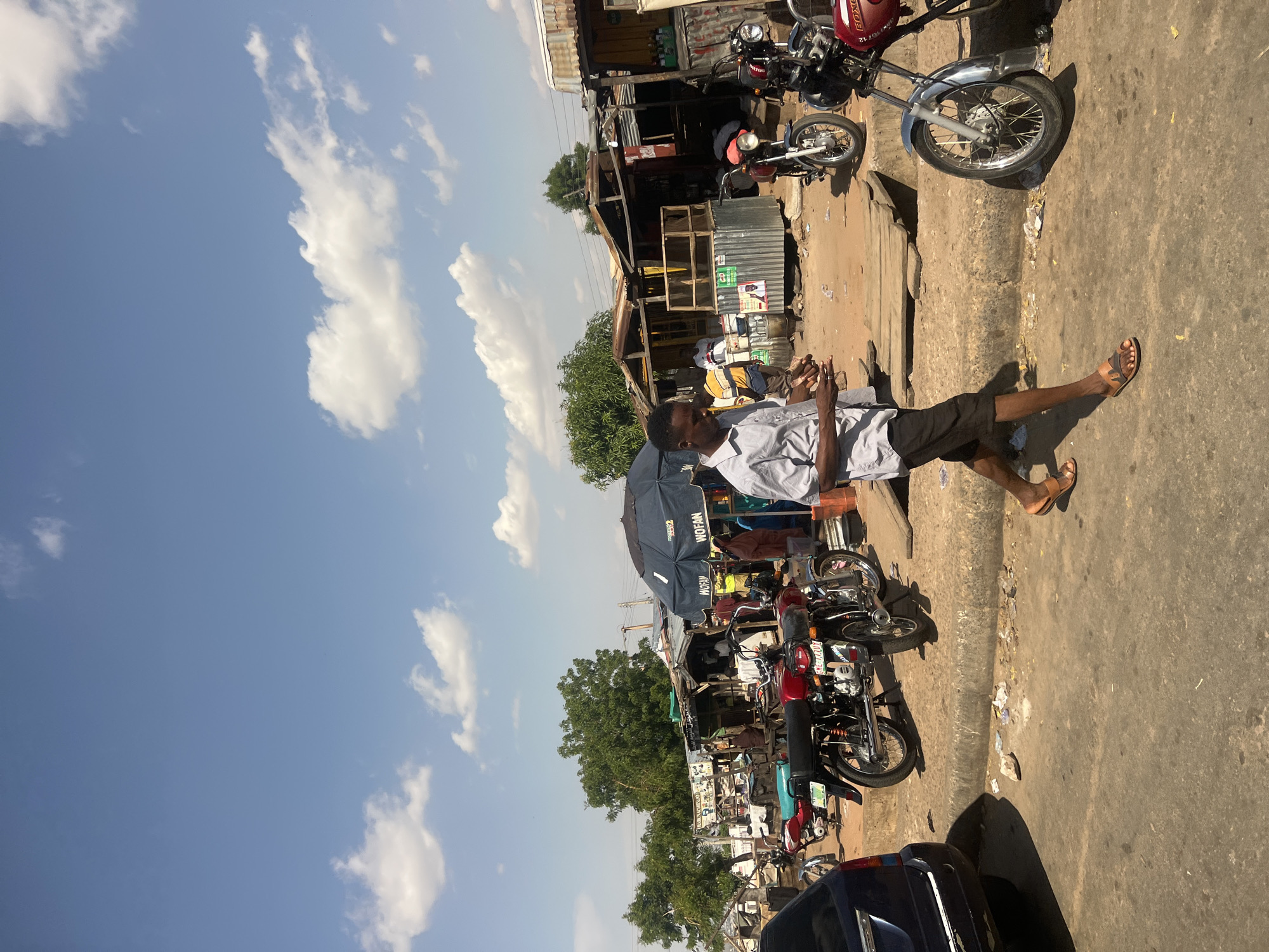 This image depicts a bustling street market scene in Gombe, Nigeria, observed under a clear blue sky with scattered white cumulus clouds. In the foreground, a paved road surface with visible dirt and scattered debris, including paper and plastic, runs along the bottom. On the far left, the rear quarter of a dark blue vehicle is partially visible. A curb separates the paved road from a raised, unpaved dirt area.
Dominating the mid-ground and extending into the foreground, a dark-skinned male is walking from left to right across the paved area, slightly angled towards the viewer. He wears a light-colored, short-sleeved collared shirt, dark shorts, and brown sandals. His gaze is directed downwards at his hands, which appear to be holding a small stack of paper currency or documents.
To the man's right and extending across the mid-ground, numerous motorcycles, primarily red and black, are parked on the dirt patch. Several are identifiable as "BOXER" brand motorcycles. Beyond these vehicles, a series of informal market stalls and shops are constructed from corrugated metal sheets, wood, and other rudimentary materials, forming a continuous line of commercial activity. Several individuals are visible within and around these stalls, engaged in various market activities. A prominent large, dark blue umbrella, branded with "WOFAN 2," provides shade over one of the stalls on the right side. Green trees are visible behind the market structures, and additional distant buildings or structures can be faintly discerned on the horizon. The scene is illuminated by strong sunlight, suggesting a clear day.