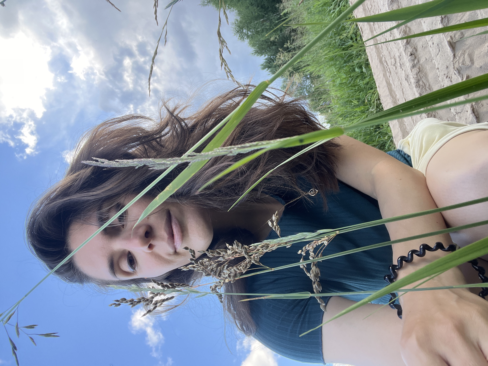 A medium shot captures a woman with dark brown hair, looking downwards and slightly to her left. She is positioned in the foreground, partially obscured by tall blades of grass and seed stalks. She wears a dark green ribbed t-shirt and light-colored shorts. A black coiled bracelet is visible on her left wrist. A delicate gold necklace with a small pendant hangs around her neck. Behind her, out of focus, is a grassy field, a line of trees, and a bright blue sky with scattered white clouds. The ground she is sitting on appears to be a sandy or gravelly embankment. The overall setting suggests an outdoor environment, possibly a park or a natural area.