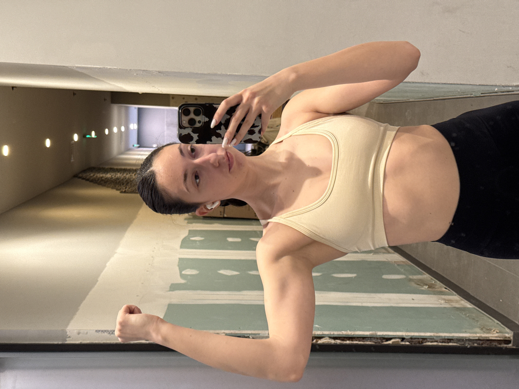 A young adult female is depicted in a mirror selfie taken within a corridor located in Sofia, Bulgaria. She has dark hair styled away from her face and is wearing a light beige sports bra and black leggings. White wireless earbuds are inserted into both ears. With her right hand, she holds a smartphone, identifiable by its black and white cow-print case, towards the mirror. Her left arm is flexed, presenting a prominent bicep. She gazes directly into the mirror/phone with a neutral expression. The immediate background behind her left arm shows a wall under renovation or construction, displaying sections of light-green drywall or primer over unfinished surfaces. The wall to her right, where the mirror is positioned, is a plain, light-colored surface. The ceiling features a series of recessed lights, some emitting warm white or yellow light, and at least two emitting green light. The floor is a light grey or beige material, and a textured wall section is partially visible further down the corridor in the reflection.