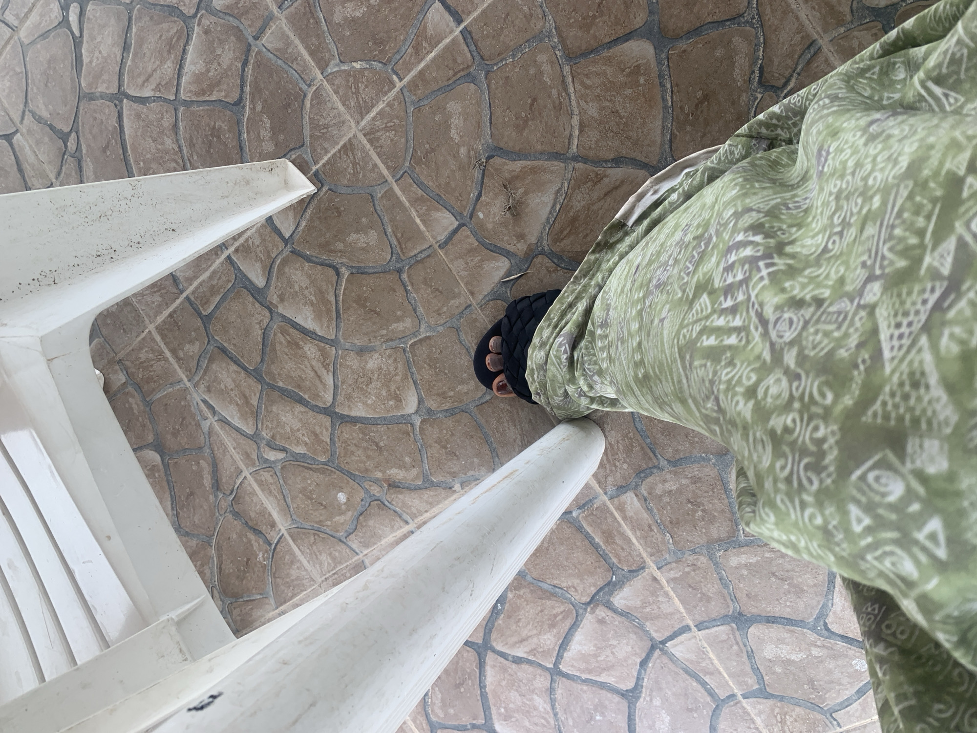 An over-the-shoulder, close-up shot shows a person's legs and feet wearing black sandals, standing on a patterned tile floor. The person is wearing a green garment with a white pattern. Part of a white plastic chair is visible in the upper left corner of the frame. The scene appears to be outdoors, possibly in a patio or courtyard area, with the tiled floor suggesting an urban setting. Given the information provided, the location is Jalingo, Nigeria.