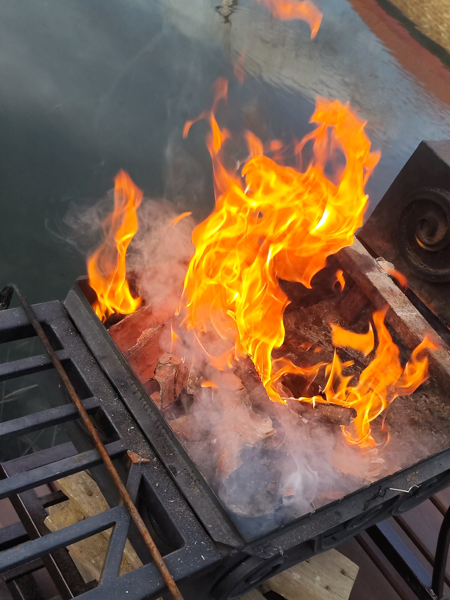 A grill situated near a body of water is ablaze with a bright orange fire. Pieces of wood are visible within the grill, and smoke rises from the burning material. The grill itself is black, with ornate metalwork on one side. The water in the background appears calm and reflects the sky. The location is potentially in the city of Chaikovsky, Russia.