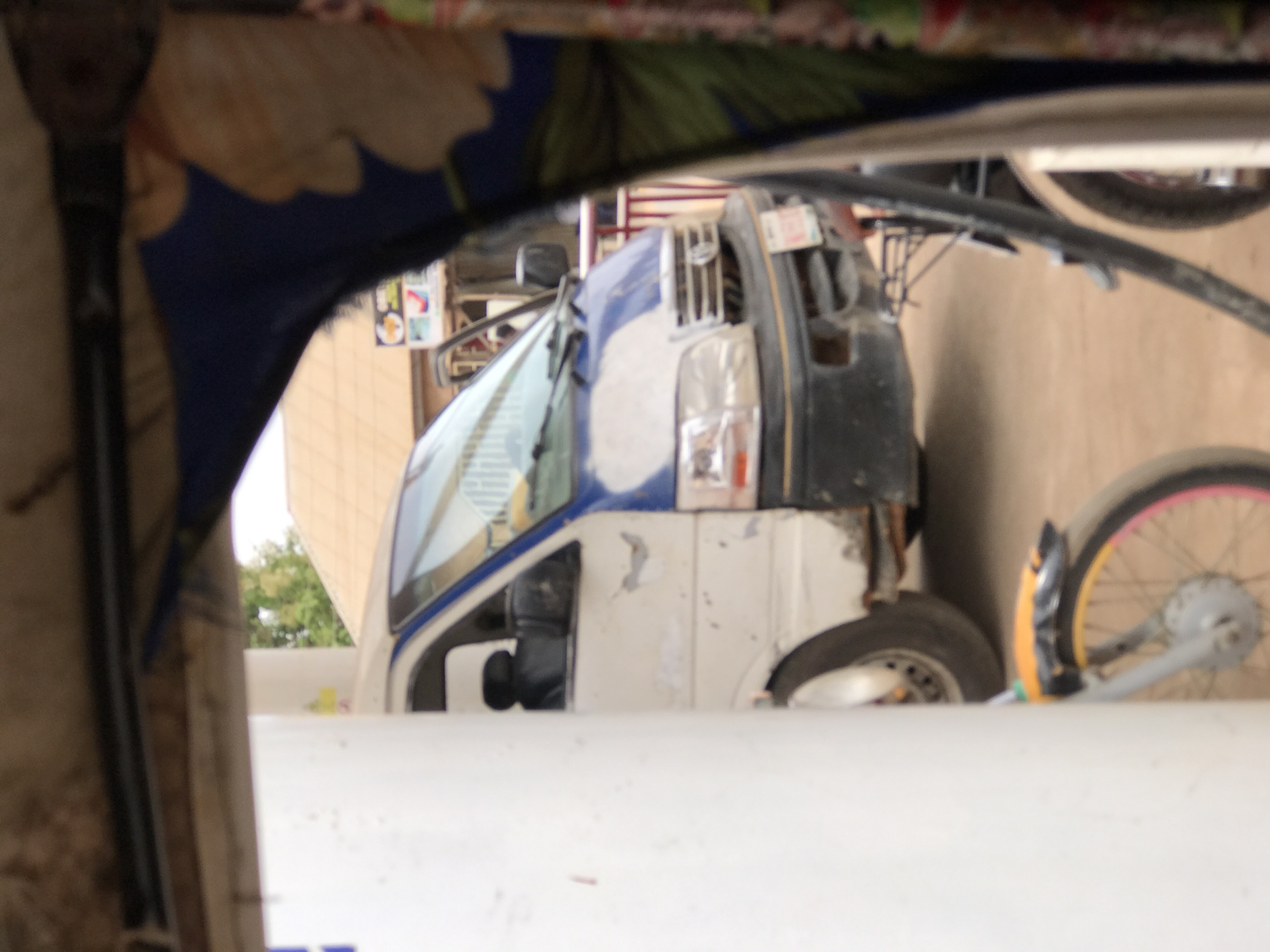 The image shows a partial view of a white and blue truck parked in an urban environment, likely in Abuja, Nigeria. The truck is seen through what appears to be the frame or part of a vehicle, obscuring part of the scene. The truck is partially obscured, revealing the front and side with a windshield and headlights. A bicycle wheel is positioned beside the truck. In the background, part of a building is visible along with some greenery.