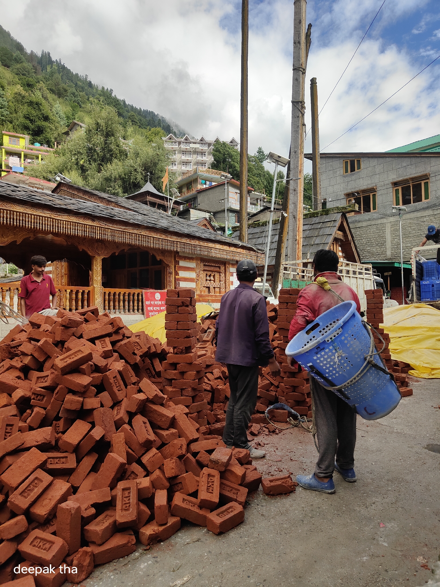 In Vashisht, India, a group of construction workers are engaged in a building project under a cloudy sky. In the foreground, a large pile of red bricks is stacked on the ground. A man in a maroon polo shirt stands to the left, observing the scene. Two other men are actively working: one in a purple shirt and grey pants, and another in a red jacket and grey pants carrying a large blue basket on his back. The background shows traditional wooden architecture with ornate carvings, typical of the region, and multi-story buildings on a forested hillside. Utility poles and wires are also visible. The overall impression is a construction site in a mountainous town, likely in the Himalayas.